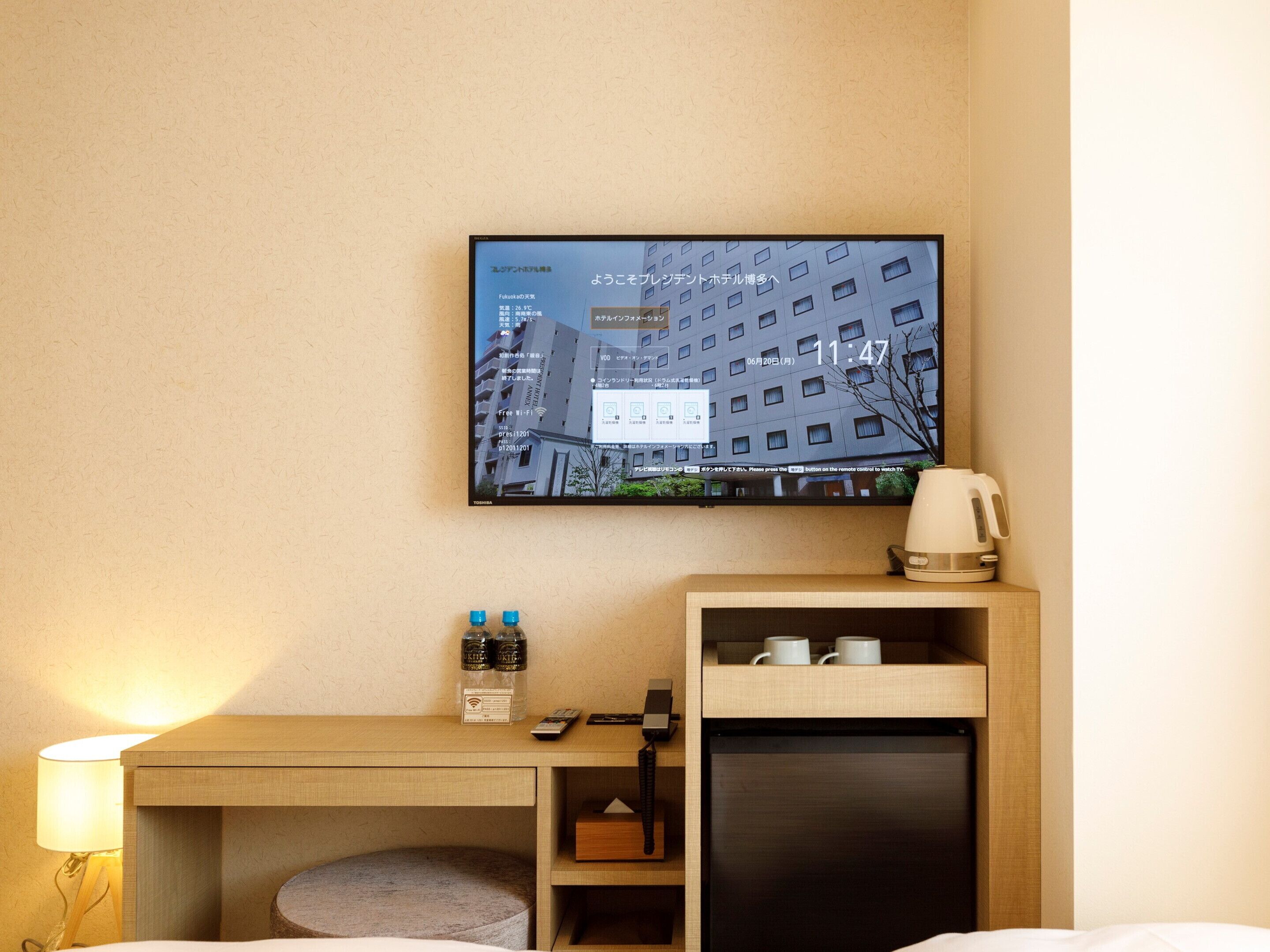 Ein Hotelzimmer mit an der Wand befestigtem Fernseher, der eine Stadtansicht zeigt, einem hölzernen Schreibtisch mit Lampe, Telefon und einer Kaffeemaschine.