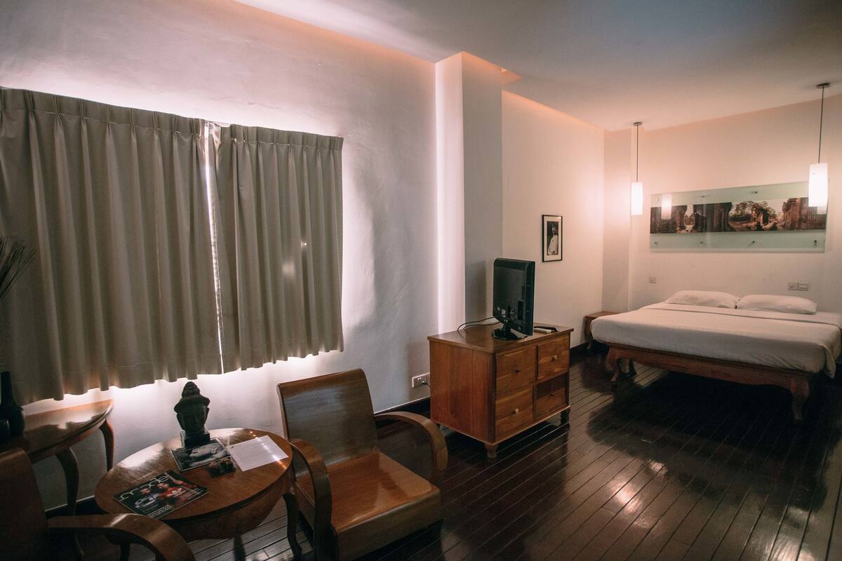 Habitación de hotel con una cama y mesita de noche de madera, televisor, escritorio con silla y ventana con cortinas.