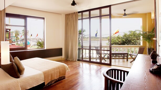 Een hotelkamer met een bed, een bureau met stoel, en een balkon met uitzicht op de straat en palmbomen.