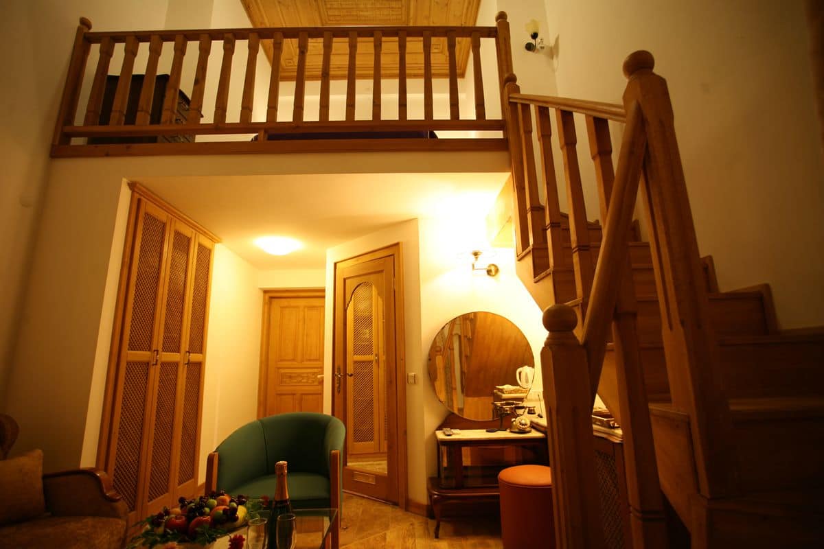 Quarto estilo loft com escada de madeira, poltrona verde, mesa pequena com abajur e porta que dá para outro quarto.