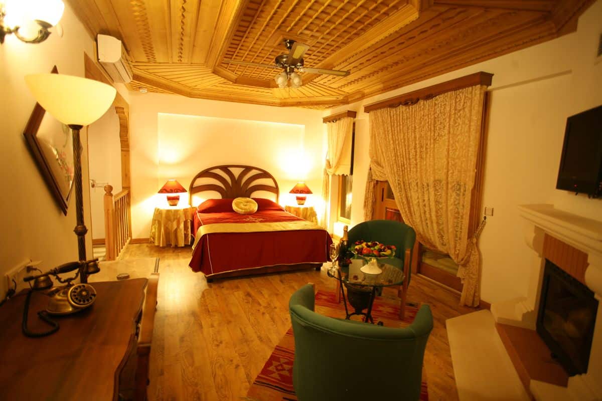 Um quarto aconchegante com teto de madeira, cama com roupa de cama vermelha, poltrona verde, lareira e telefone antigo.