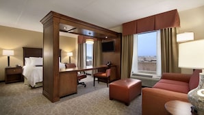 A hotel room with a bed, desk, chair, ottoman, and a large window with curtains. - Hampton Inn & Suites Charles Town (Charles Town)