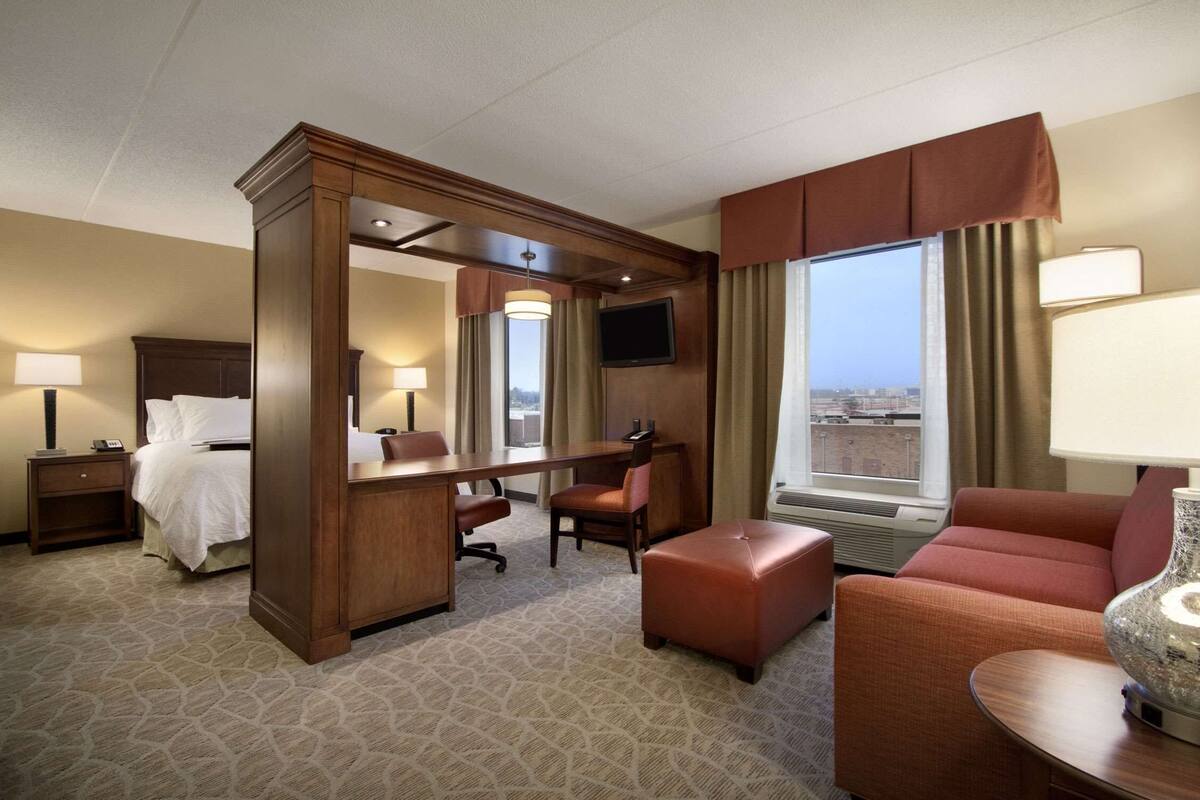 A hotel room with a bed, desk, chair, ottoman, and a large window with curtains.