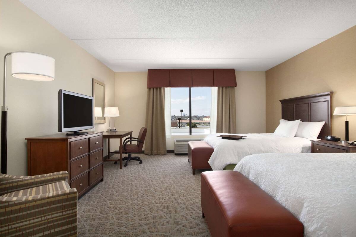 A hotel room with two beds, a desk with a computer, a TV, a chair, and a large window with a city view.