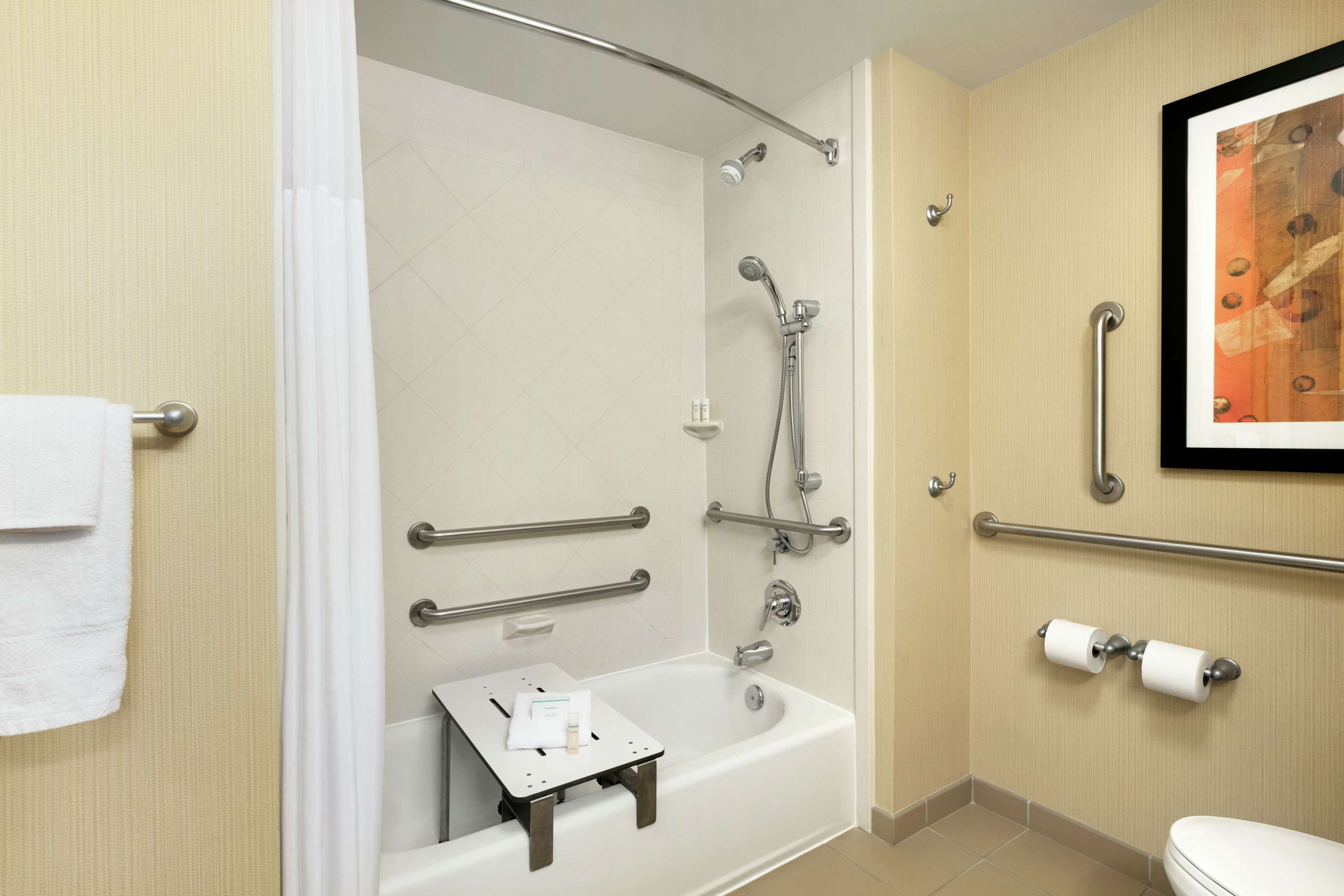 Un baño con bañera, ducha e inodoro. Cuenta con barras de apoyo, un perchero y una pequeña mesa con una toalla y una botella.