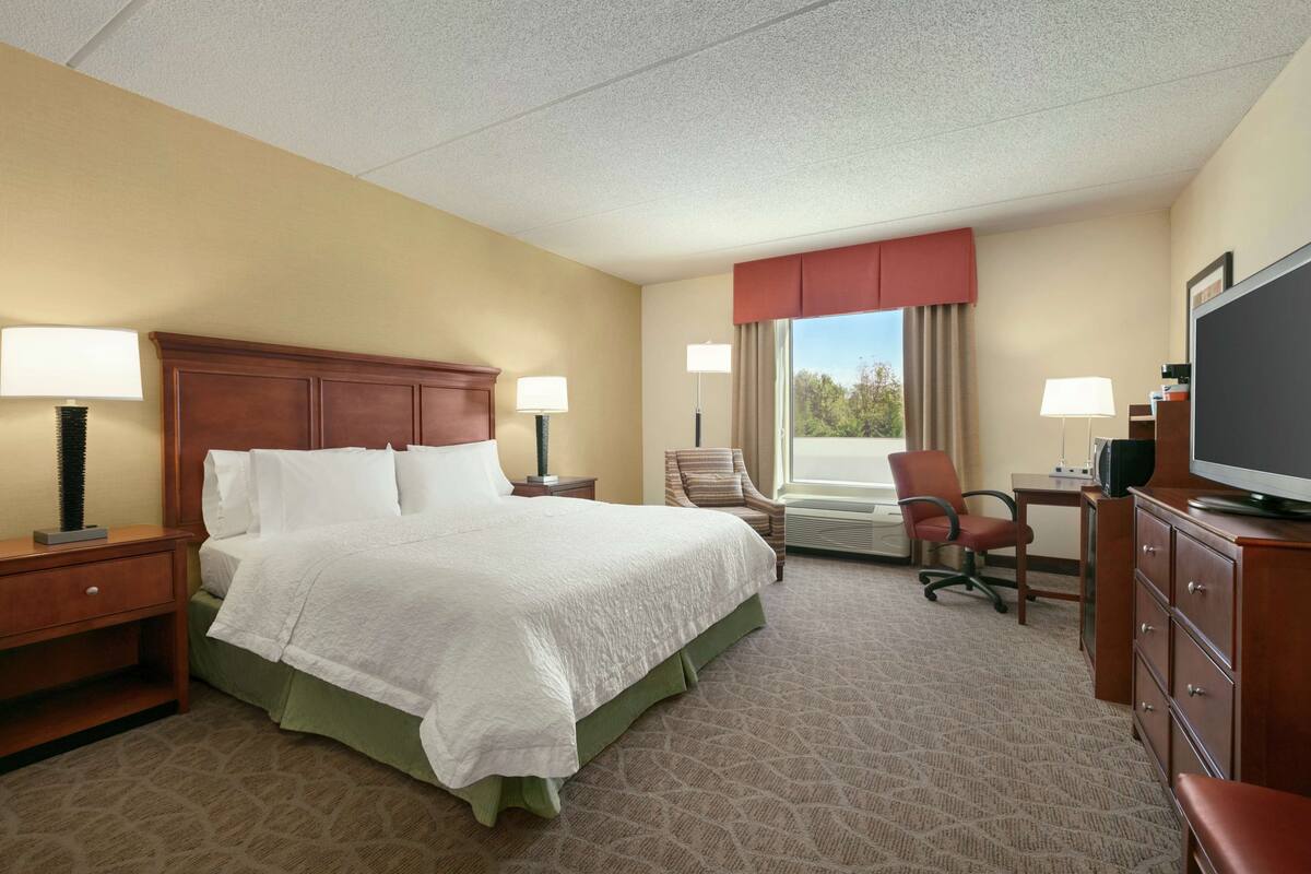 A hotel room with a large bed, two bedside tables with lamps, a desk with a chair, a television, and a window with curtains.