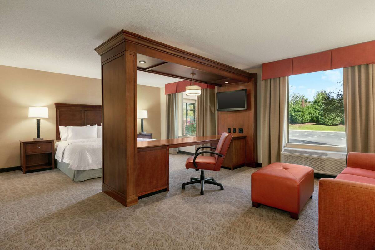 A hotel room with a bed, desk, chair, ottoman, and a large window with a view of greenery.