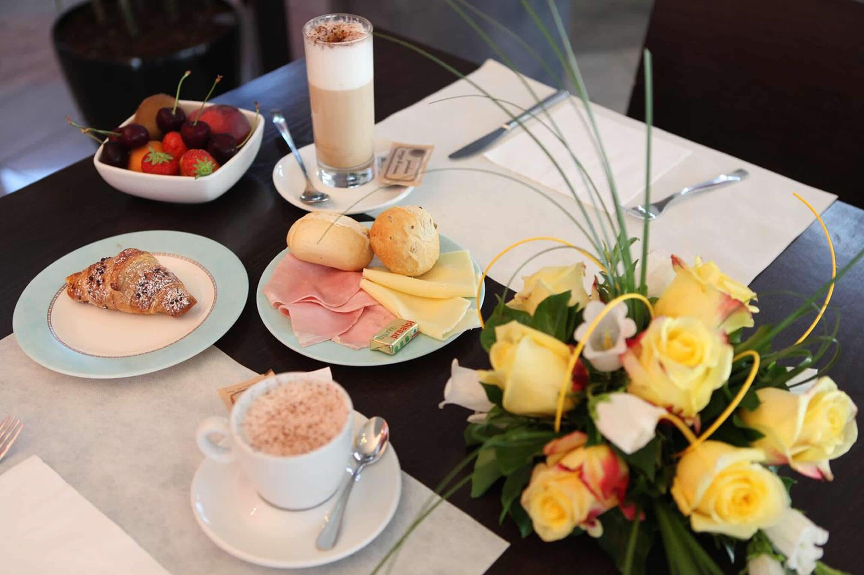 A table set with a croissant, ham and cheese, a fruit bowl, a glass of coffee, and a bouquet of yellow roses.