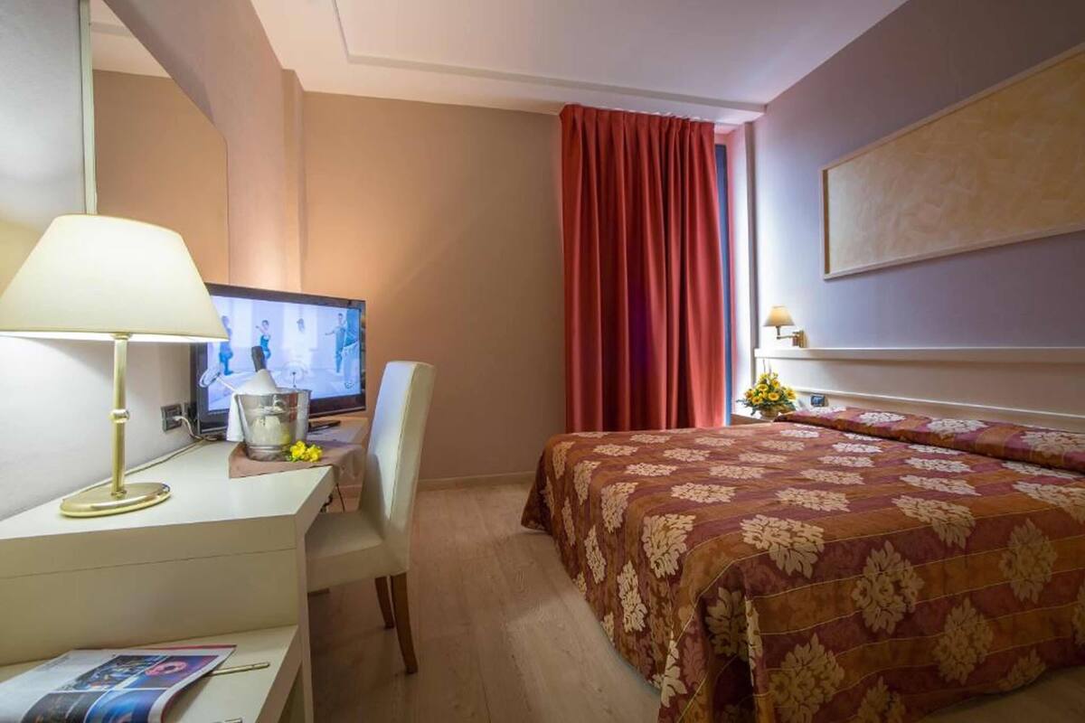 A hotel room with a bed, a desk with a lamp, a television, and a window with curtains.