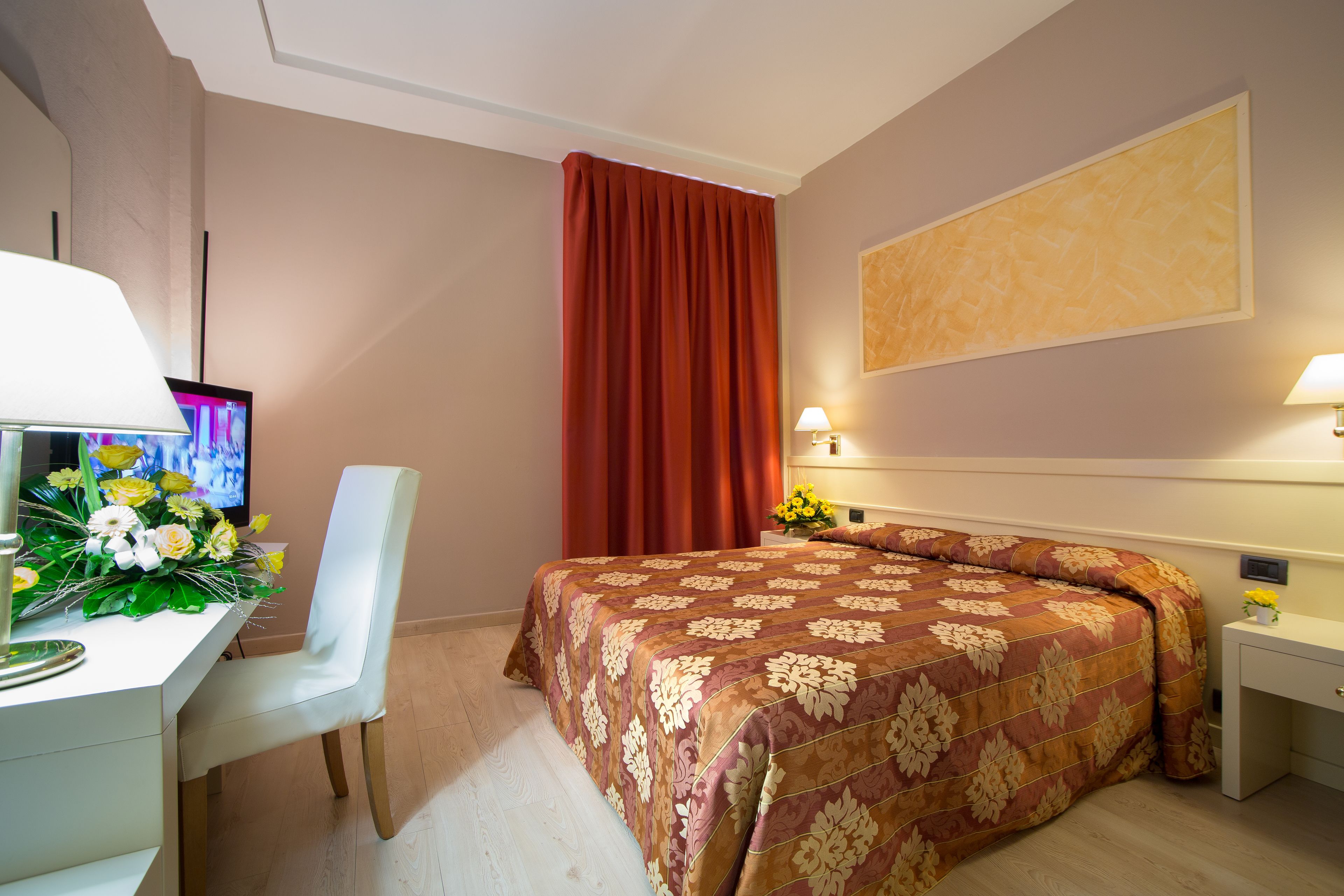 A hotel room with a bed, a desk with a computer and flowers, a TV, and a painting on the wall.