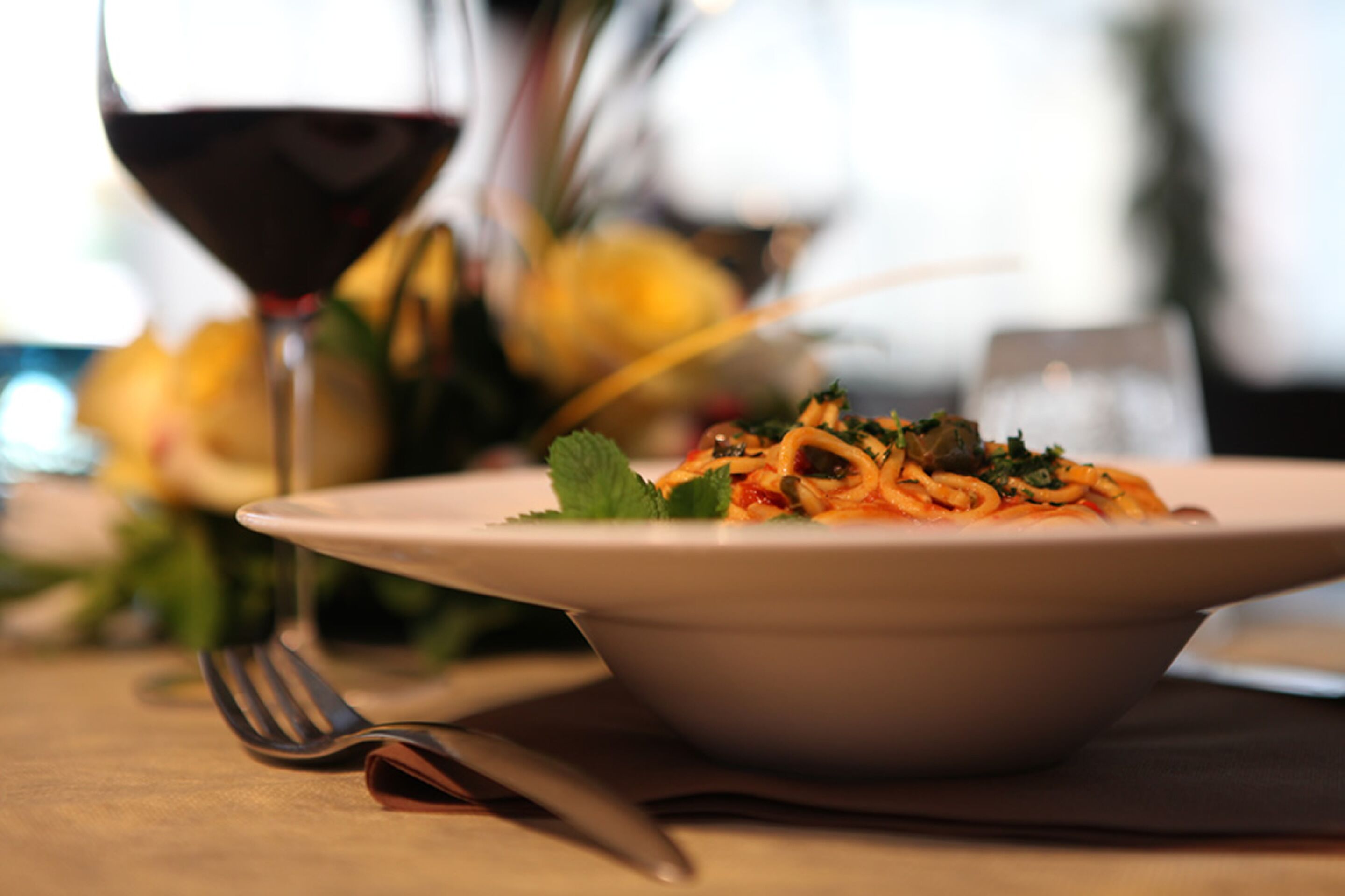 A bowl of pasta with a fork and knife on a table, with a glass of red wine in the background.
