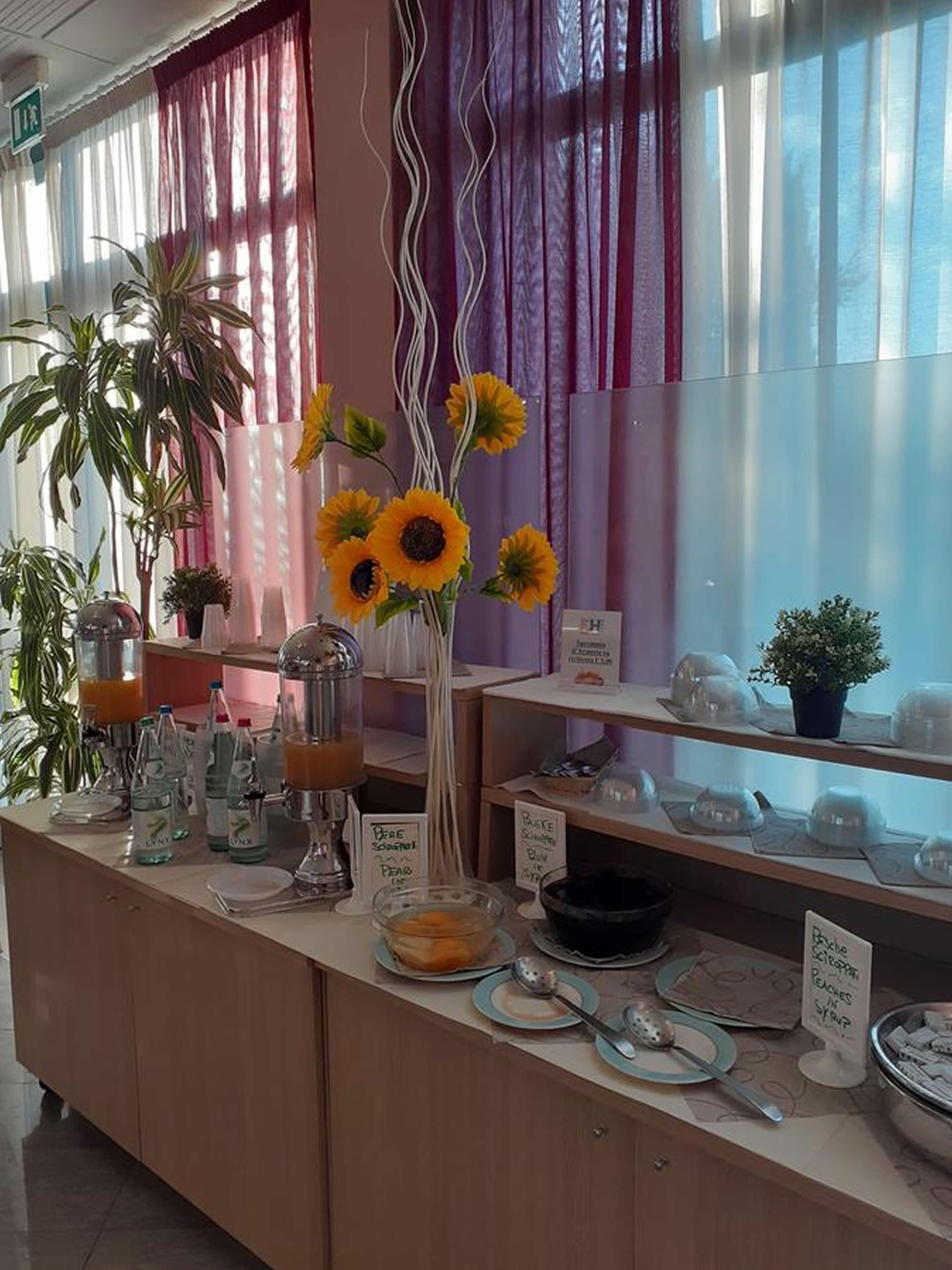 Ein Frühstücksbuffet mit einer Auswahl an Speisen und Getränken, darunter eine große Vase mit Sonnenblumen, eine Kaffeemaschine und mehrere Schilder, die die angebotenen Gerichte kennzeichnen.