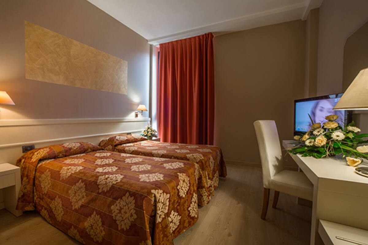 A hotel room with two beds, a desk with a computer, a chair, and a window with curtains.
