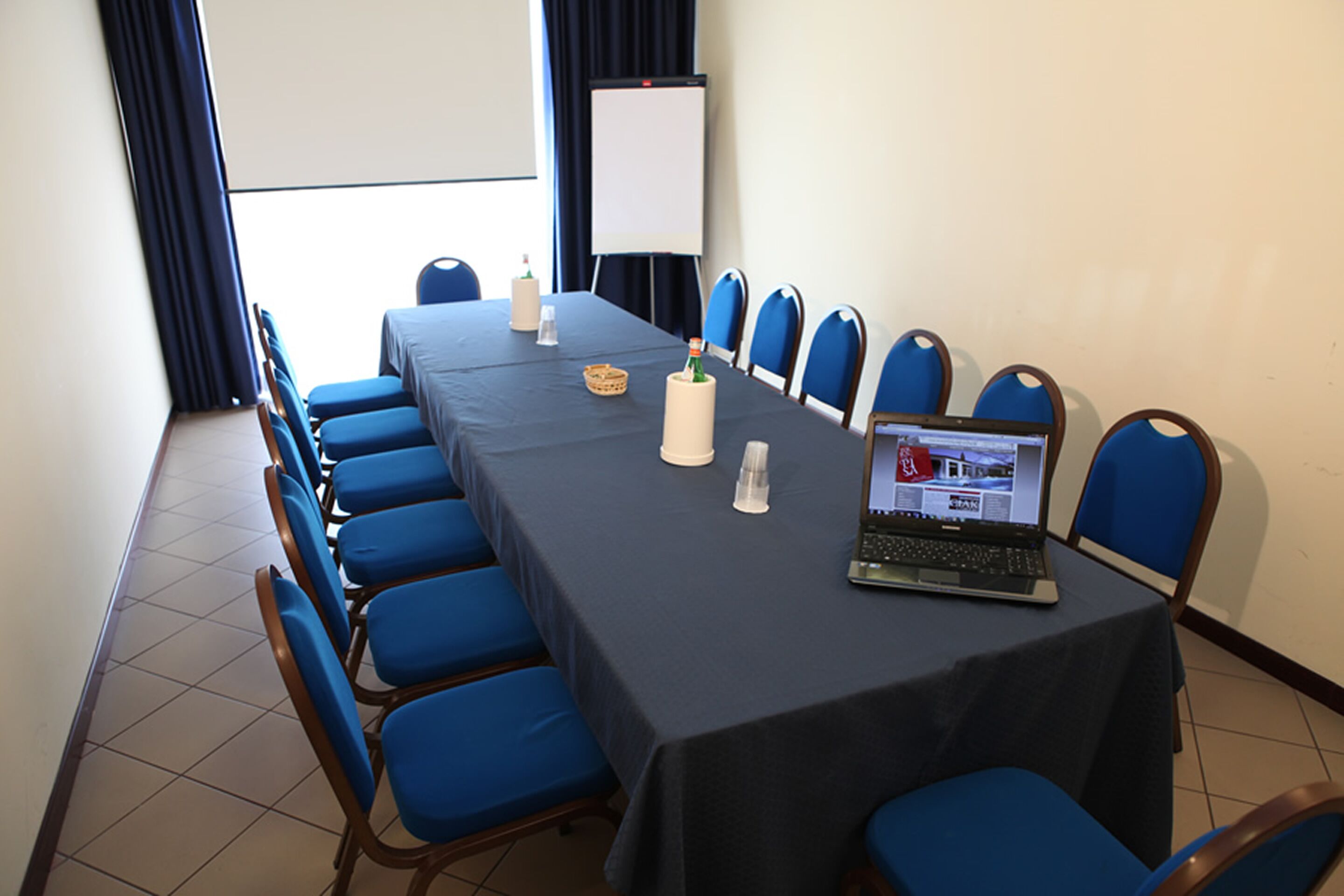 A conference room with a long table, blue chairs, a laptop, a whiteboard, and a projector screen.
