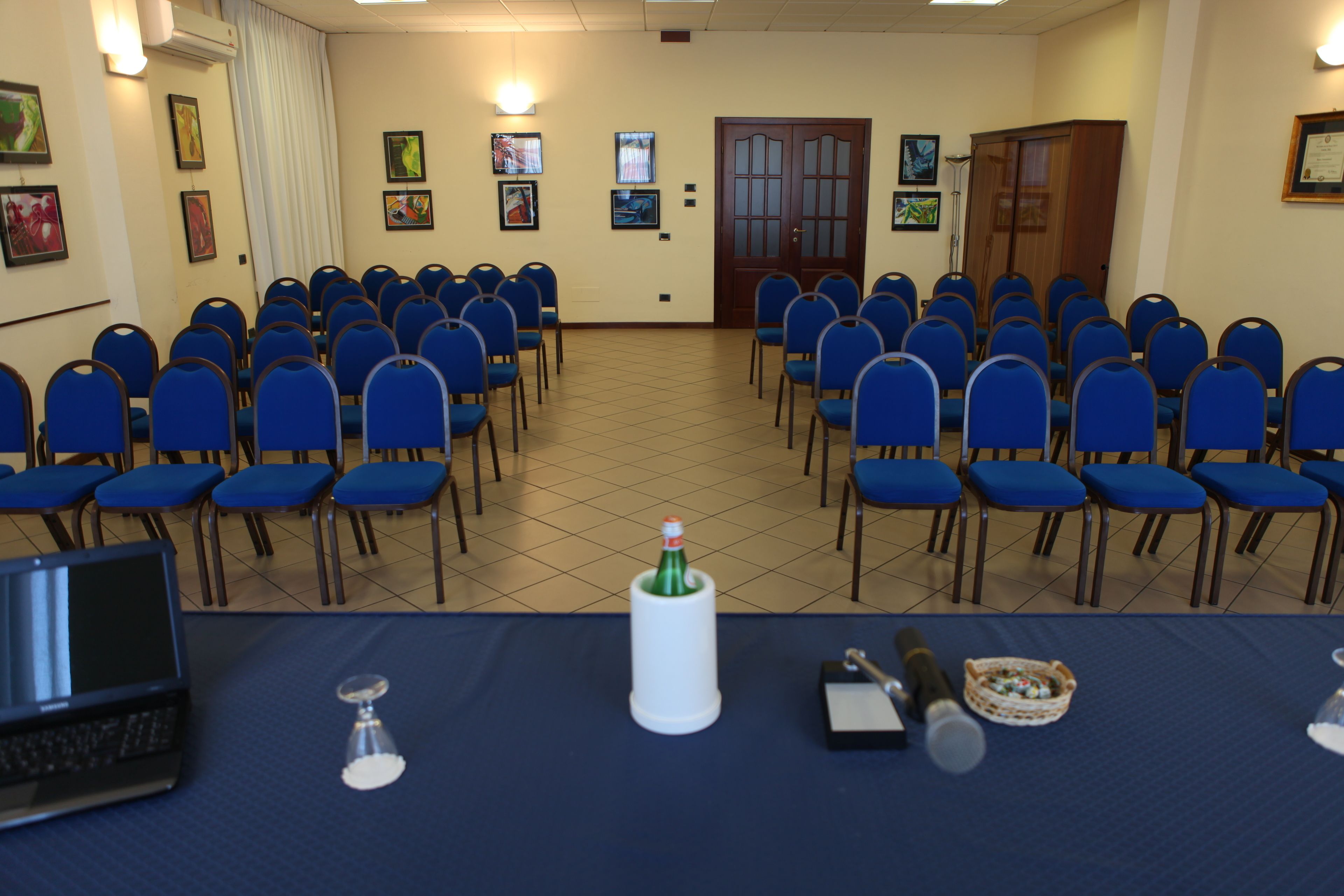 Une salle de conférence avec des rangées de chaises bleues, un ordinateur portable, une bouteille et un panier sur une table.