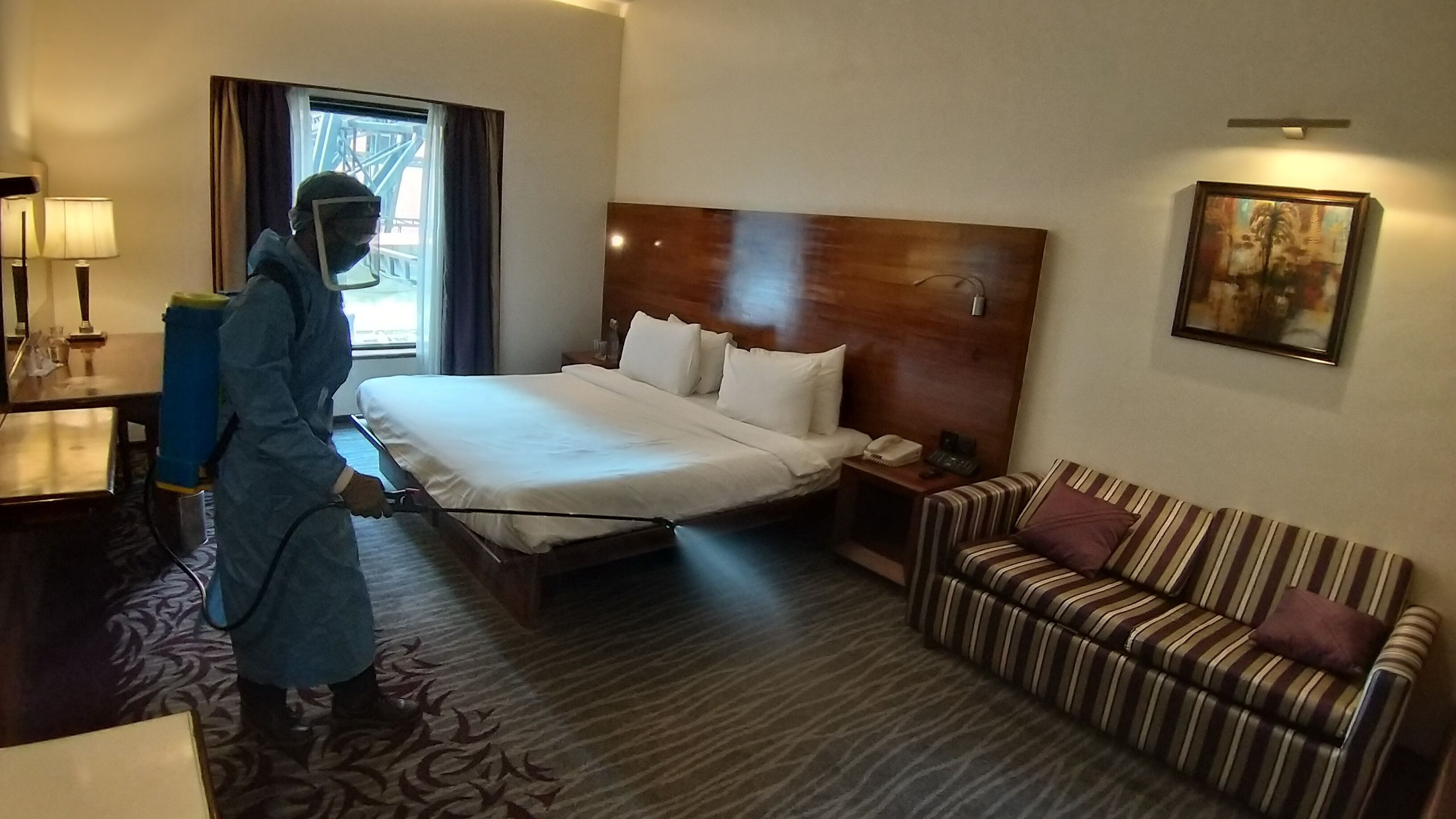 A hotel room with a bed, a striped sofa, a nightstand, a lamp, and a painting on the wall.