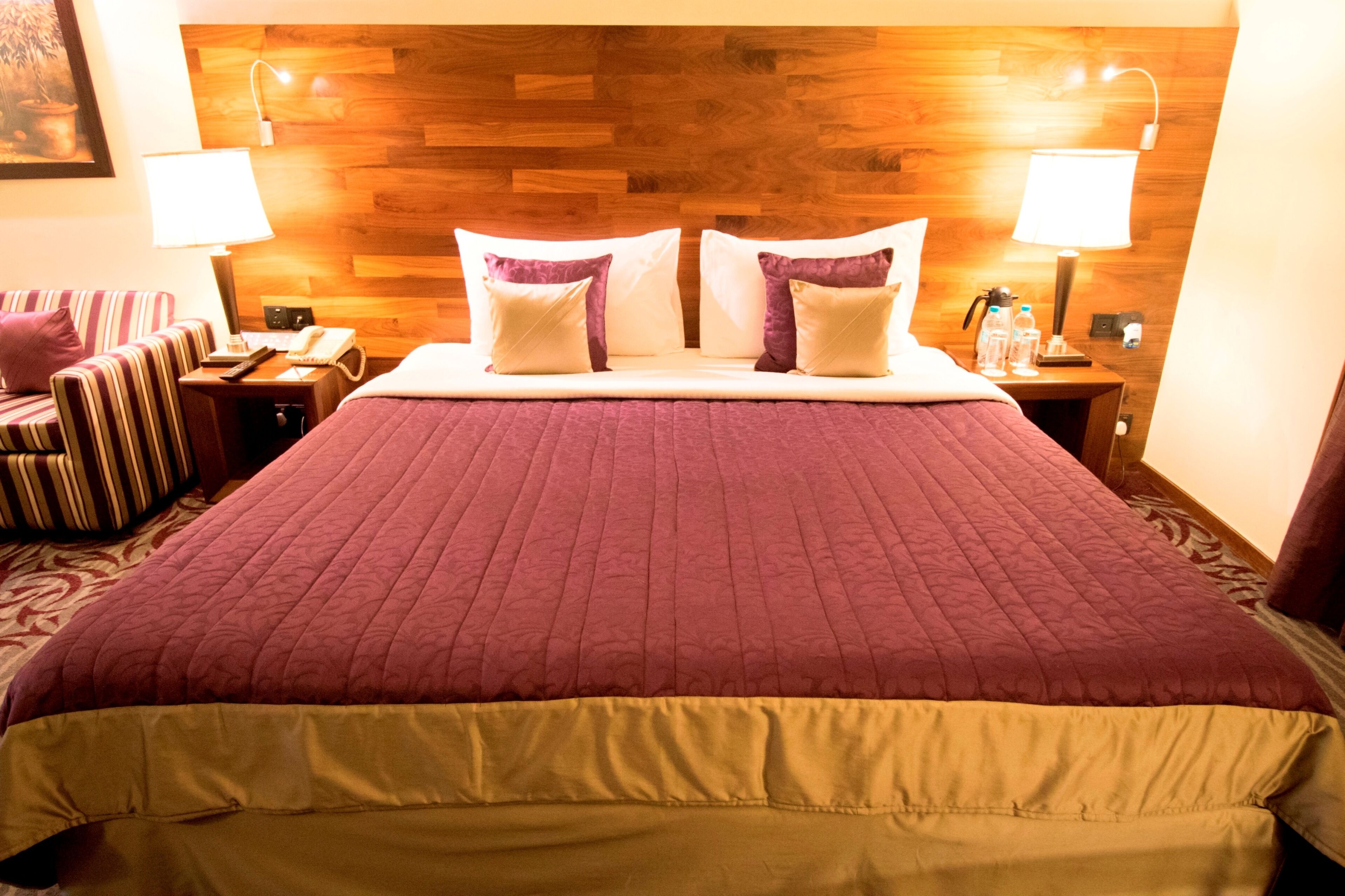 A hotel room with a large bed, two bedside lamps, a nightstand with a phone and a lamp, a striped chair, and a wooden headboard.