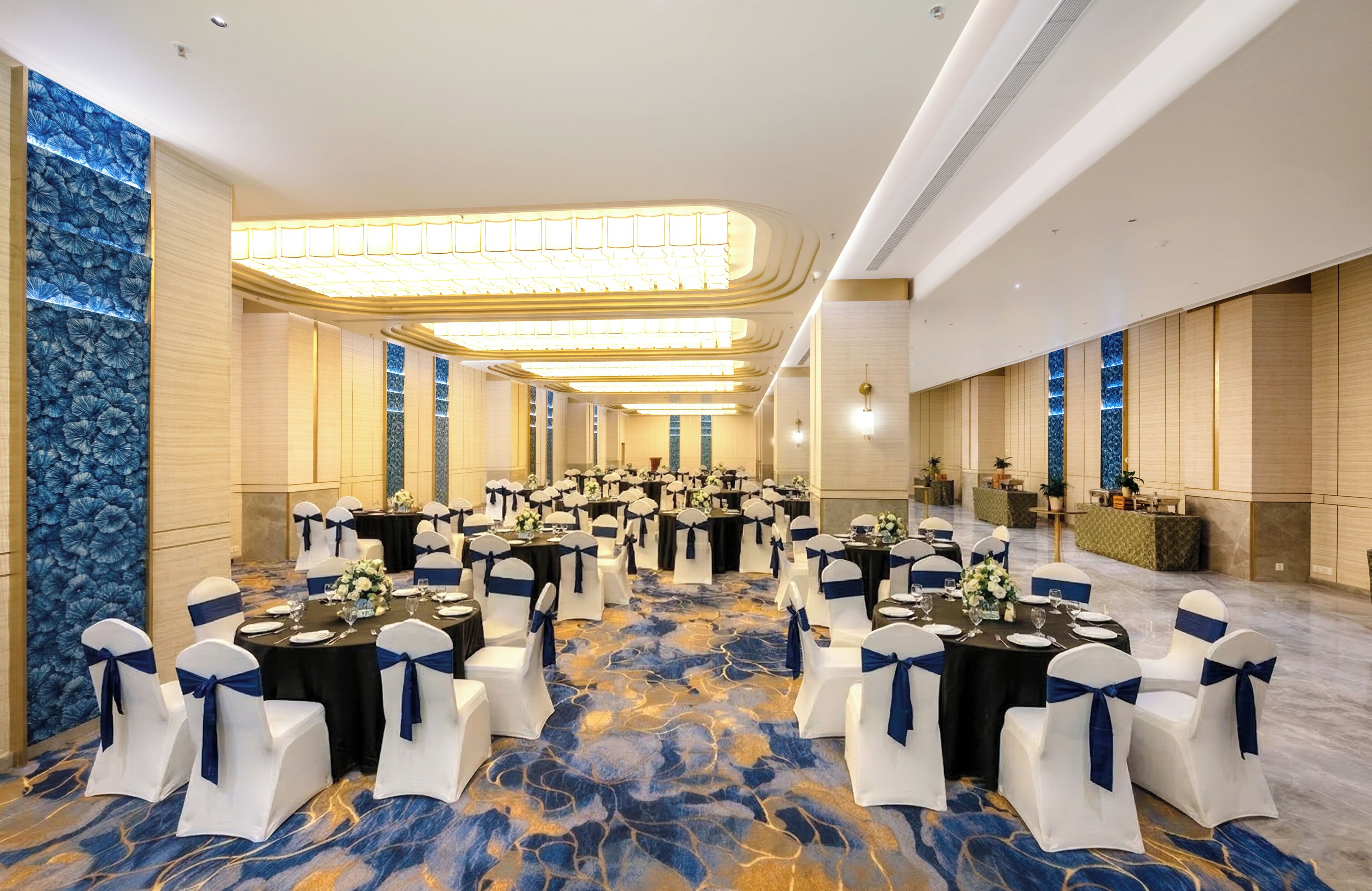 Un amplio salón de banquetes con filas de mesas y sillas, una alfombra estampada y un techo alto con iluminación empotrada.