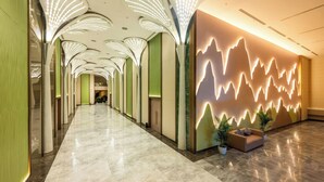 Un vestíbulo de hotel con un largo pasillo, lámparas modernas, un área de descanso con plantas y una pared con un diseño de onda luminosa.
