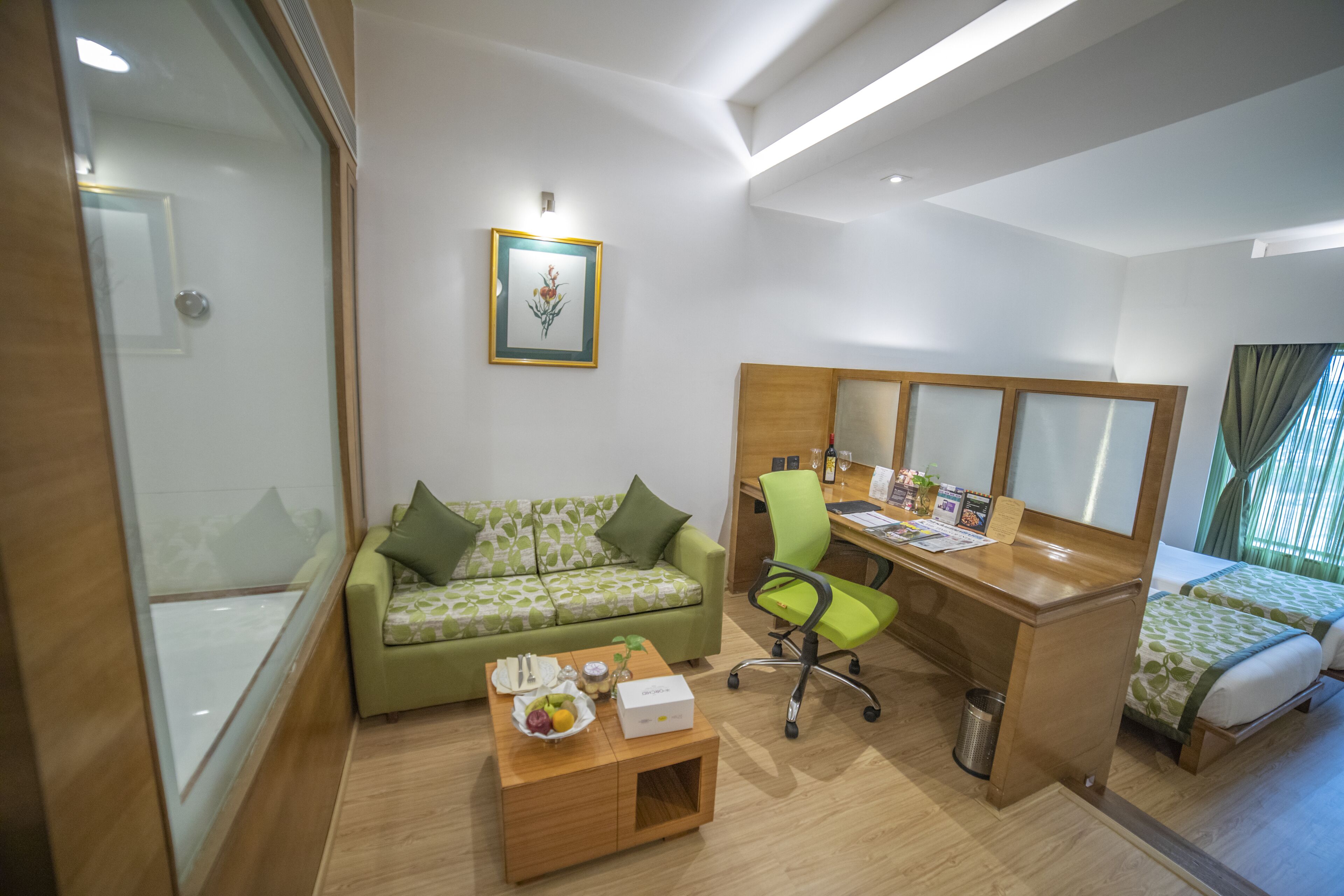 A hotel room with a green patterned sofa, a wooden desk, a green office chair, a small wooden side table with a fruit bowl, a framed picture on the wall, and a bed with green leaf-patterned bedding.