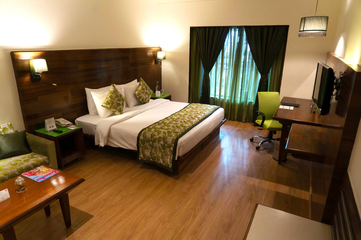 Habitación de hotel con cabecera de madera, cama con ropa blanca y detalles verdes, escritorio con silla, televisor y ventana con cortinas.