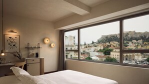 A hotel room with a large window offering a view of a historic cityscape, a neatly made bed, a small desk with a lamp, and a wall-mounted artwork. - A for Athens (Athens)
