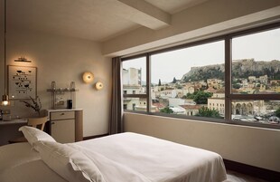 Habitación de hotel con un ventanal amplio que ofrece vistas a un casco antiguo, una cama bien hecha, un escritorio pequeño con lámpara y una obra de arte colgada en la pared.