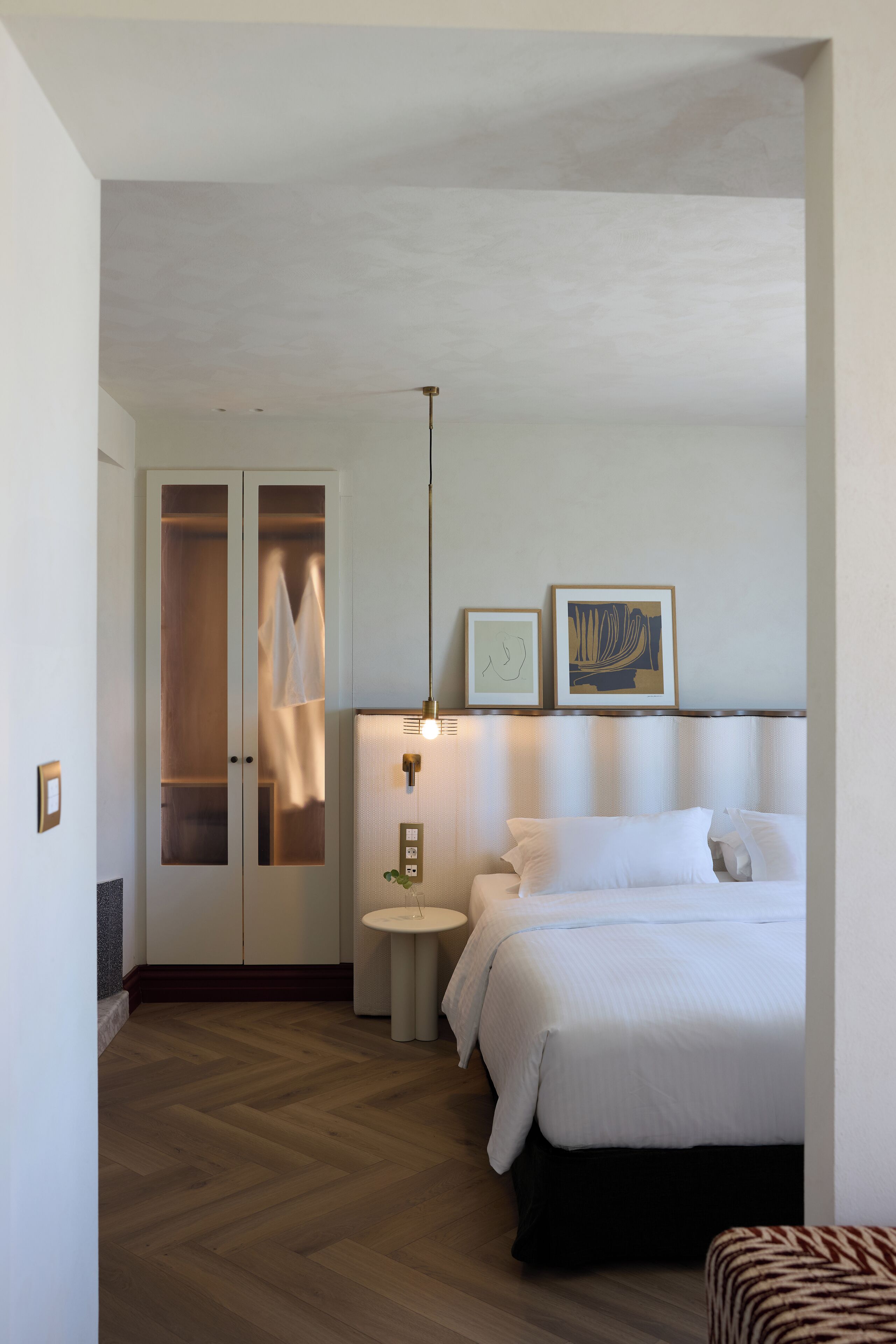 Quarto de hotel com cama, mesa de cabeceira, luminária, guarda-roupa e duas molduras na parede.
