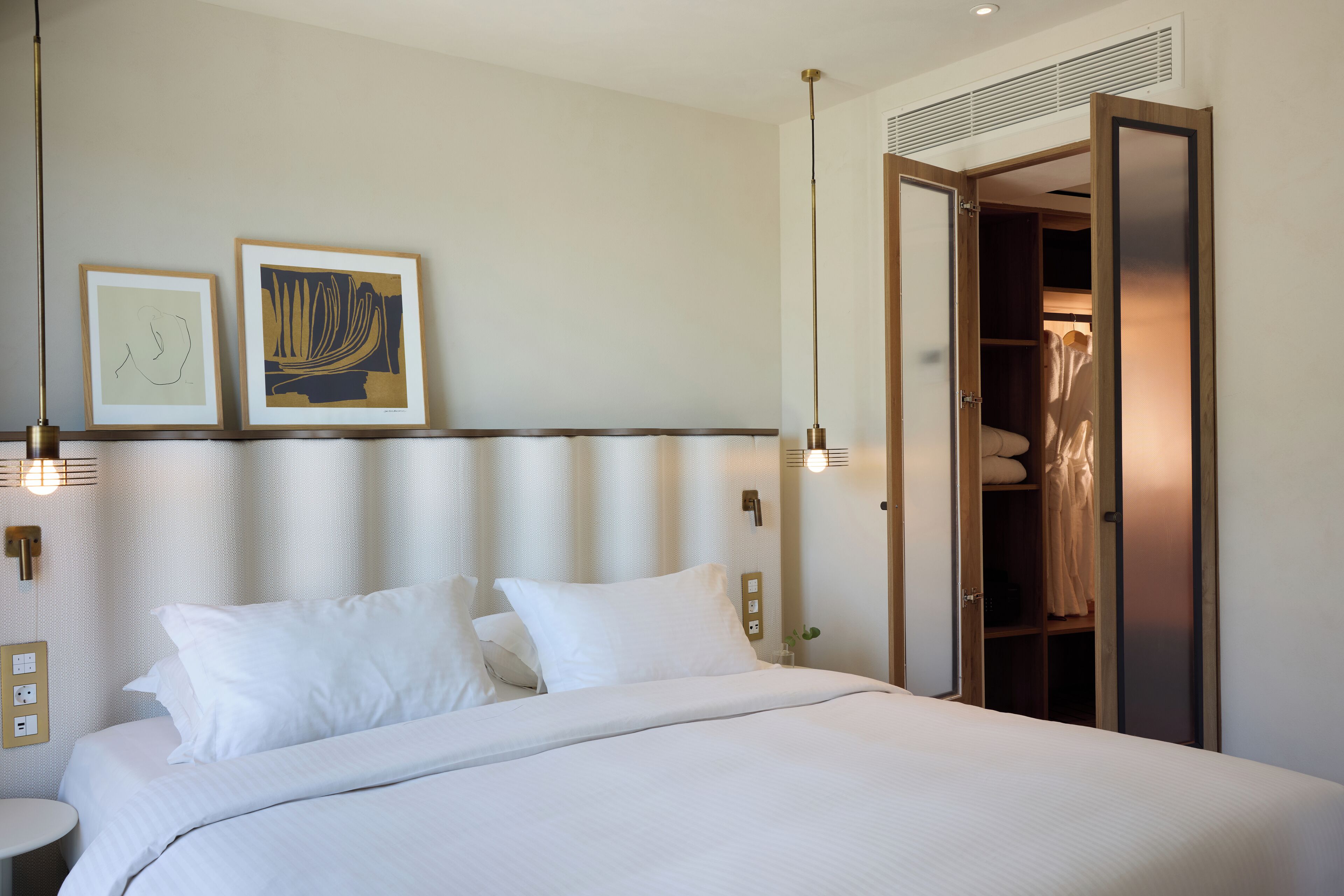 A hotel room with a neatly made bed, a bedside table, a wall-mounted light, and a wardrobe with a mirror.