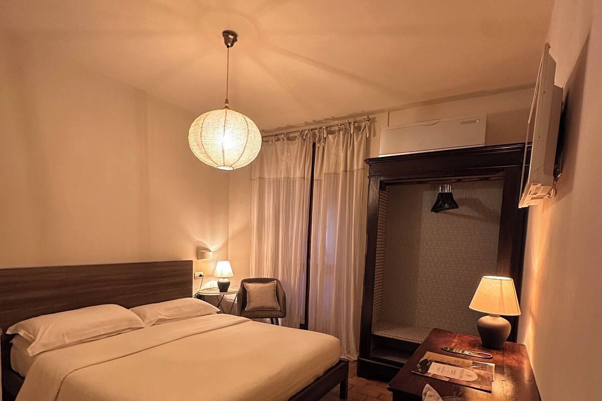 A hotel room with a double bed, a chair, a desk with a lamp, a mirror, and a hanging light fixture.