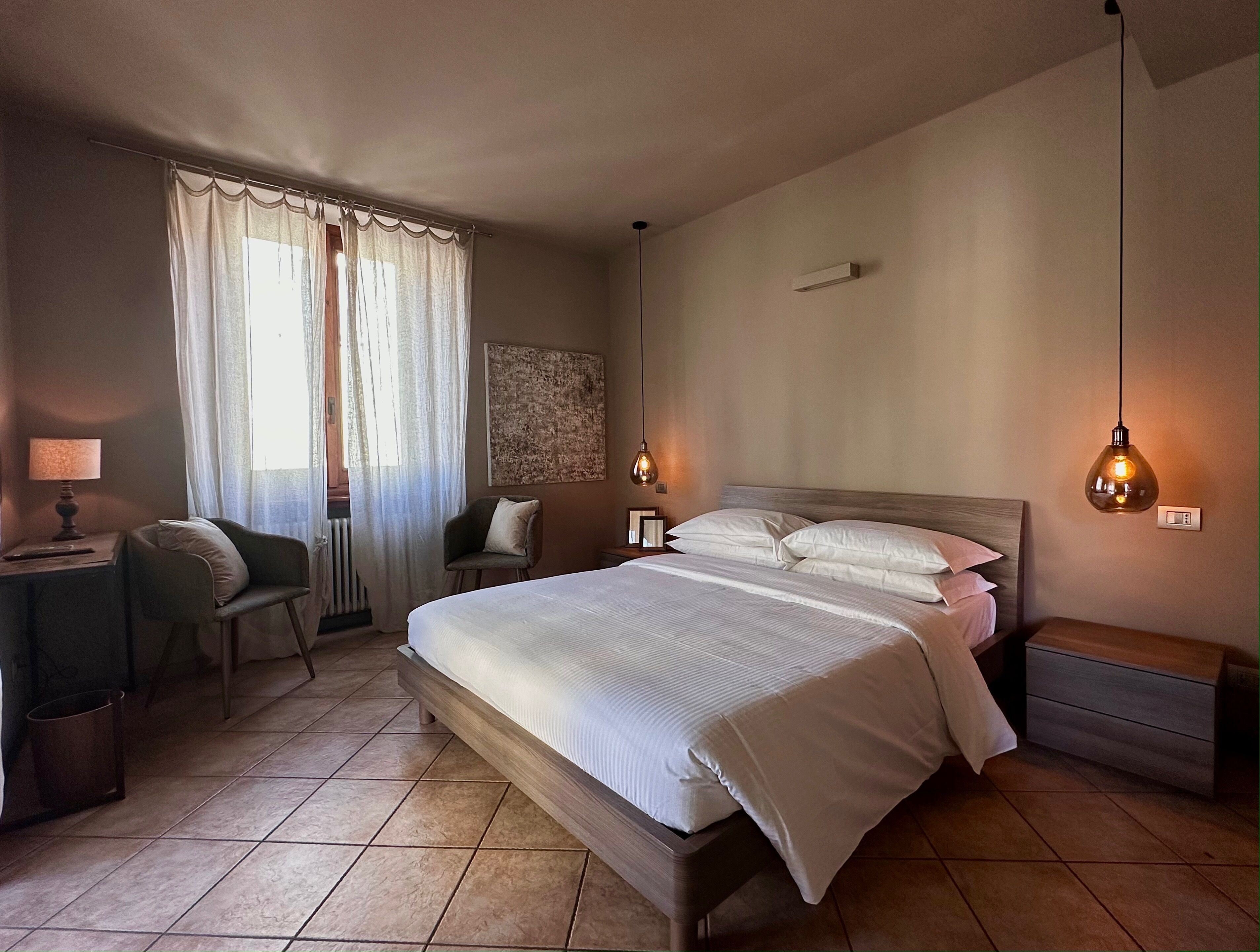Un dormitorio con cama, mesitas de noche, una silla, lámpara y ventana con cortinas.