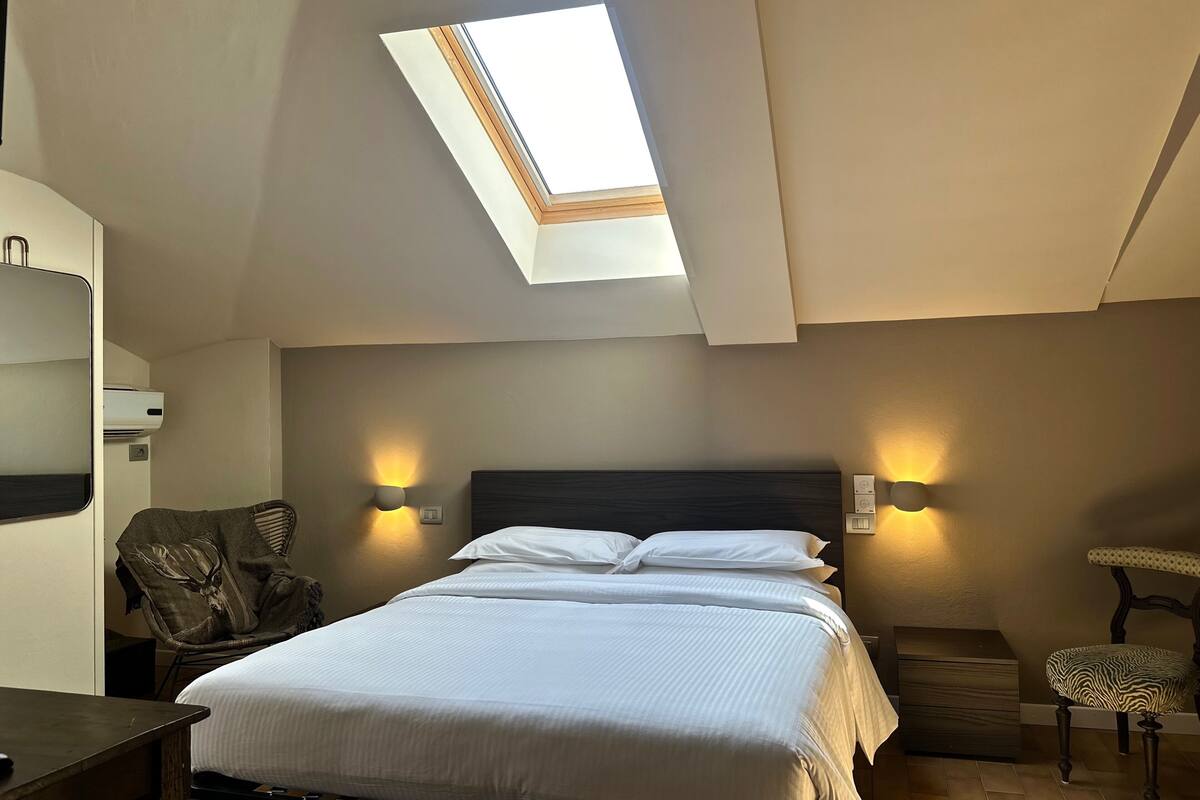 A bedroom with a skylight, a bed, two wall-mounted lamps, a chair, and a small wooden chest.