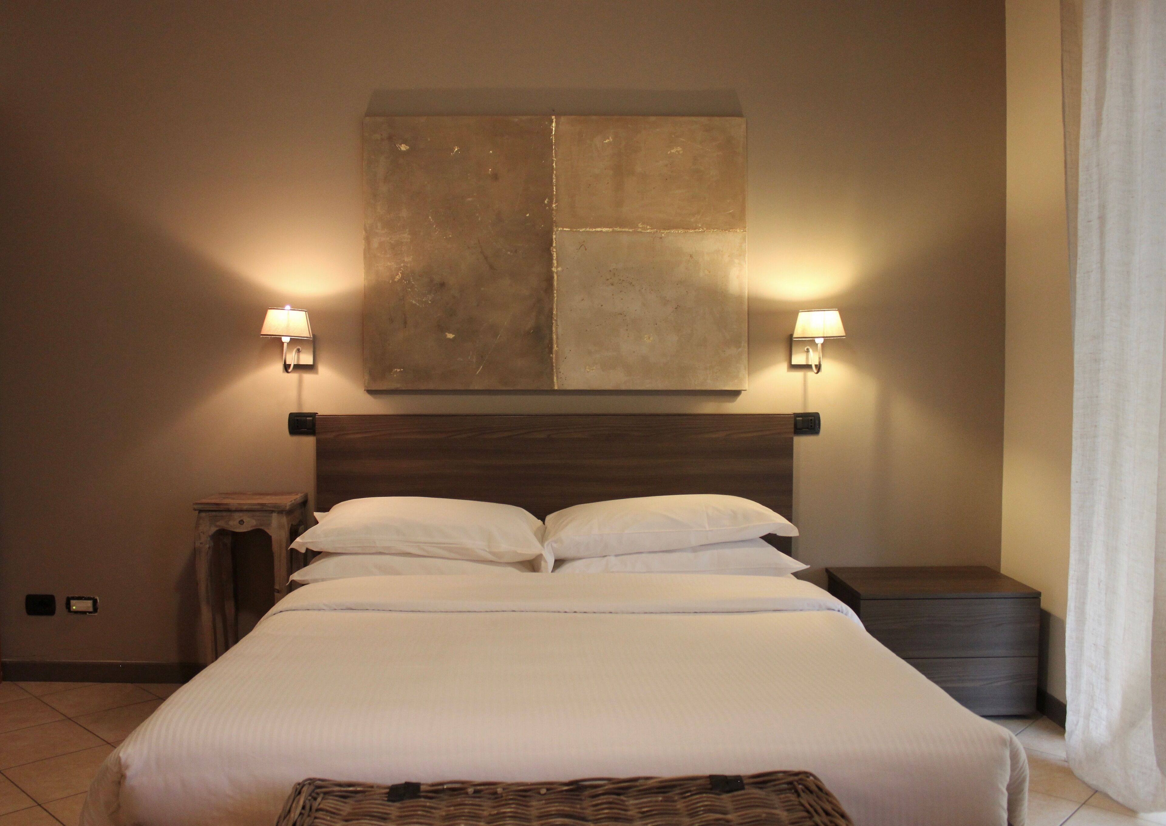 A neatly made bed with white linens, two bedside lamps, a large abstract painting above the bed, and a small wooden nightstand to the left.