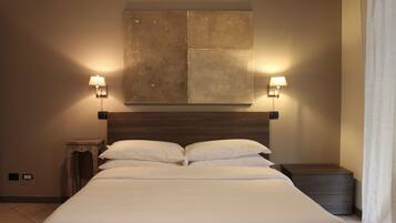 A neatly made bed with white linens, two bedside lamps, a large abstract painting above the bed, and a small wooden nightstand to the left.