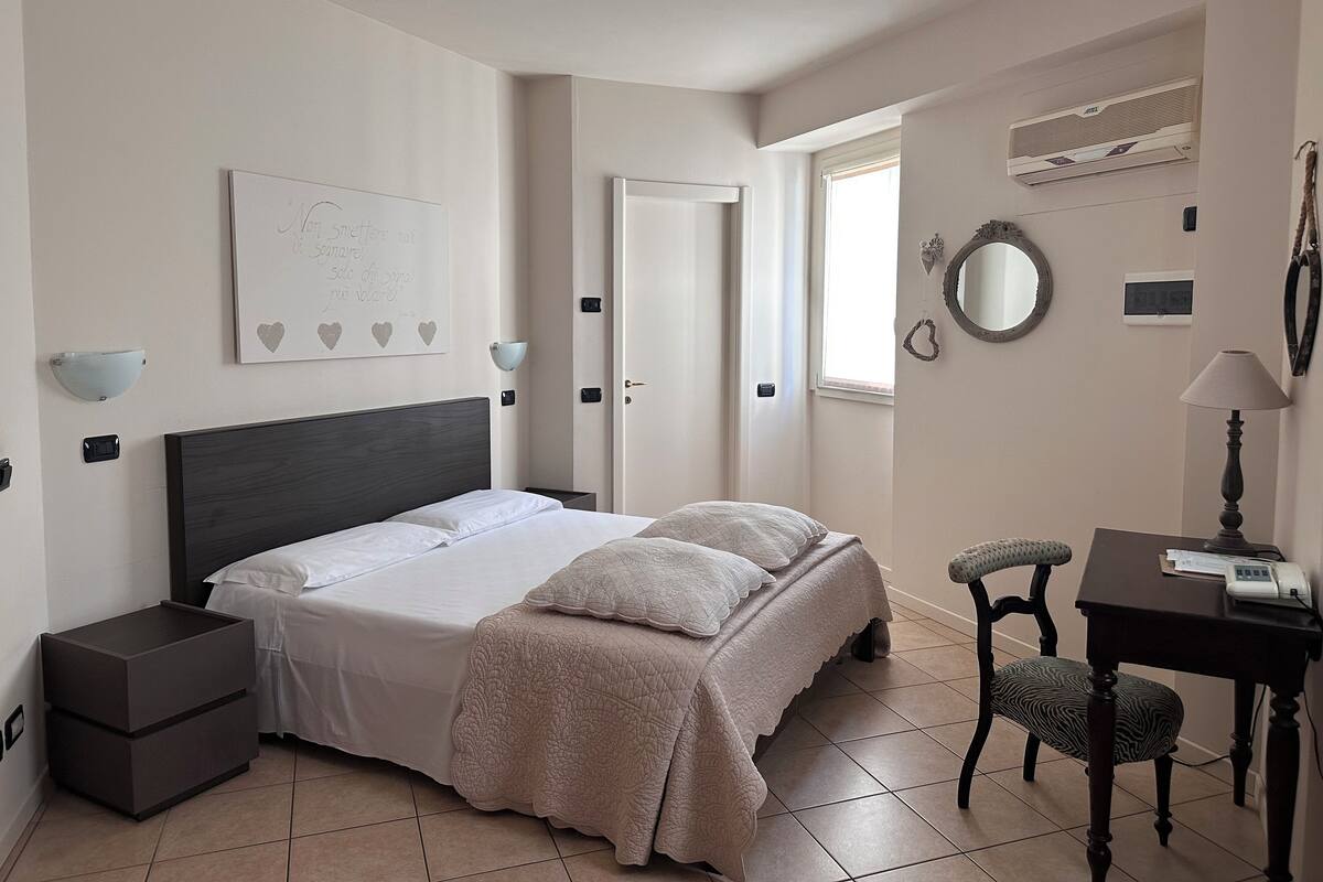 A bedroom with a bed, bedside tables, a desk with a chair, a mirror, and a wall-mounted air conditioner.