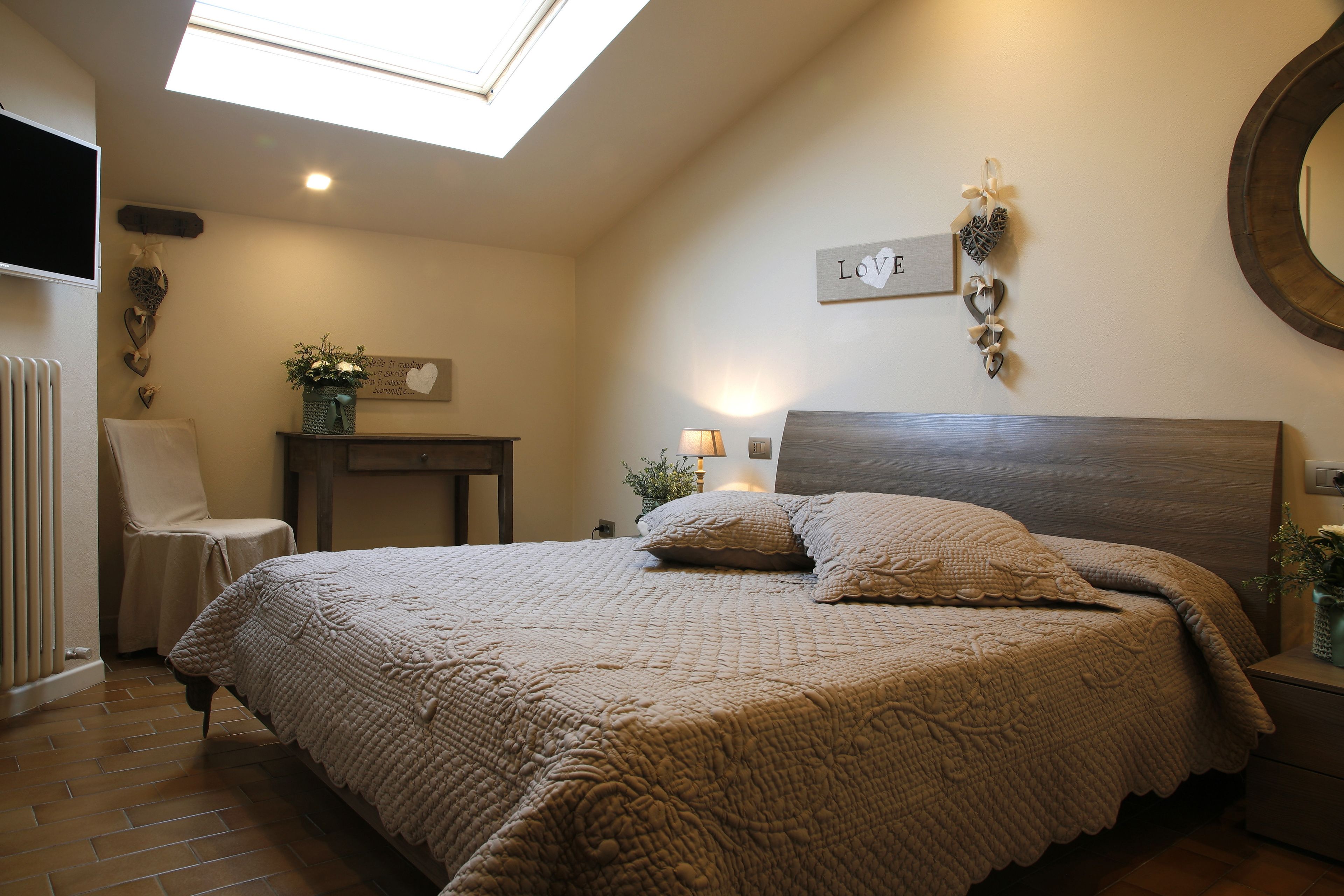 A bedroom with a large bed, a skylight, a chair, a bedside table, and a wall-mounted mirror.
