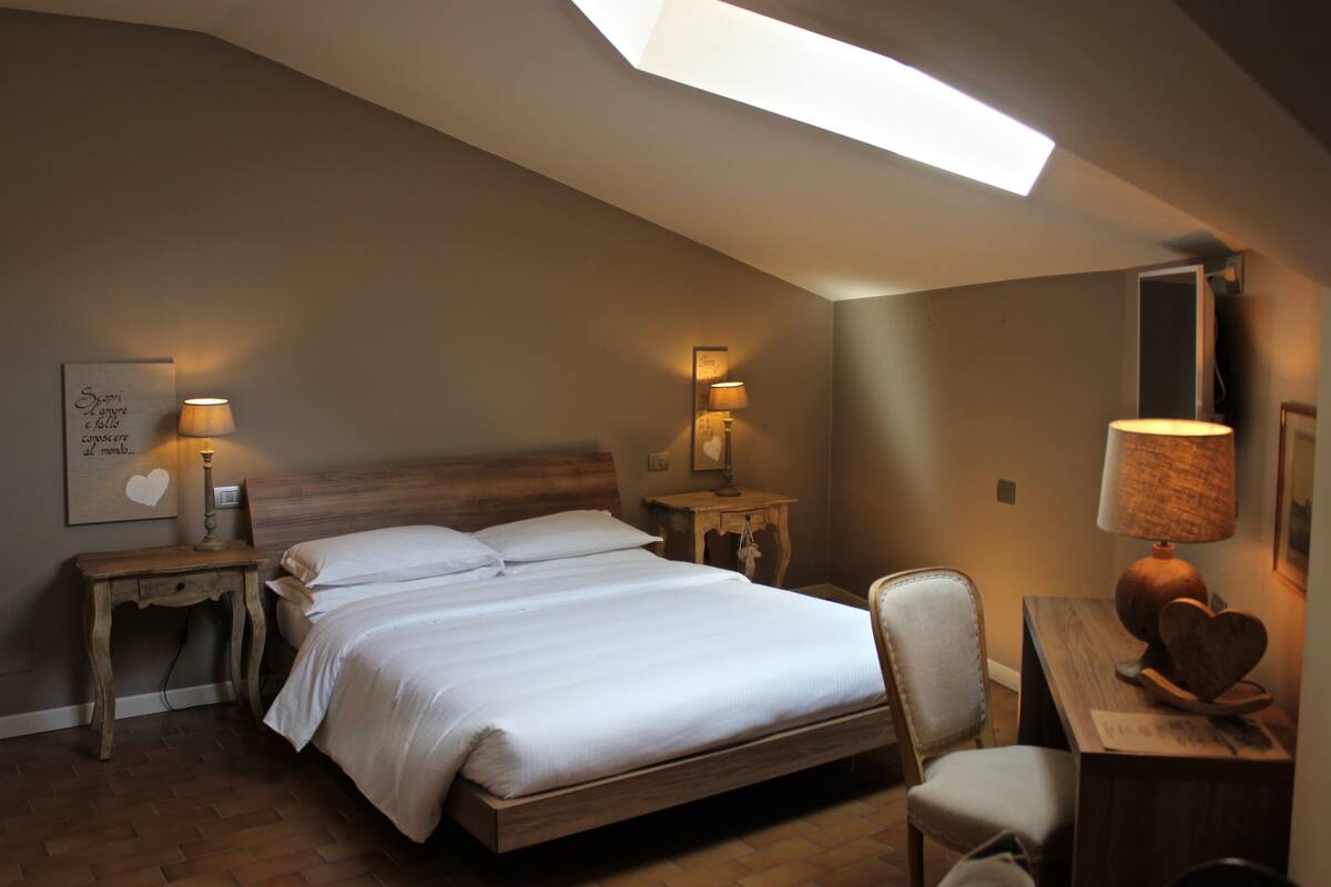 A bedroom with a skylight, a bed, two bedside tables with lamps, a chair, and a desk with a lamp.