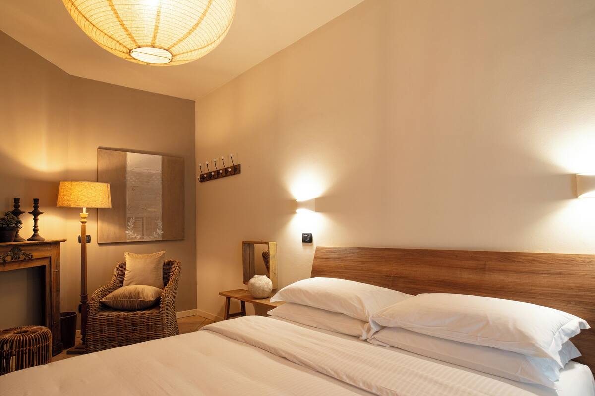 A hotel room with a bed, a chair, a lamp, a wall-mounted light, a mirror, and a wooden headboard.