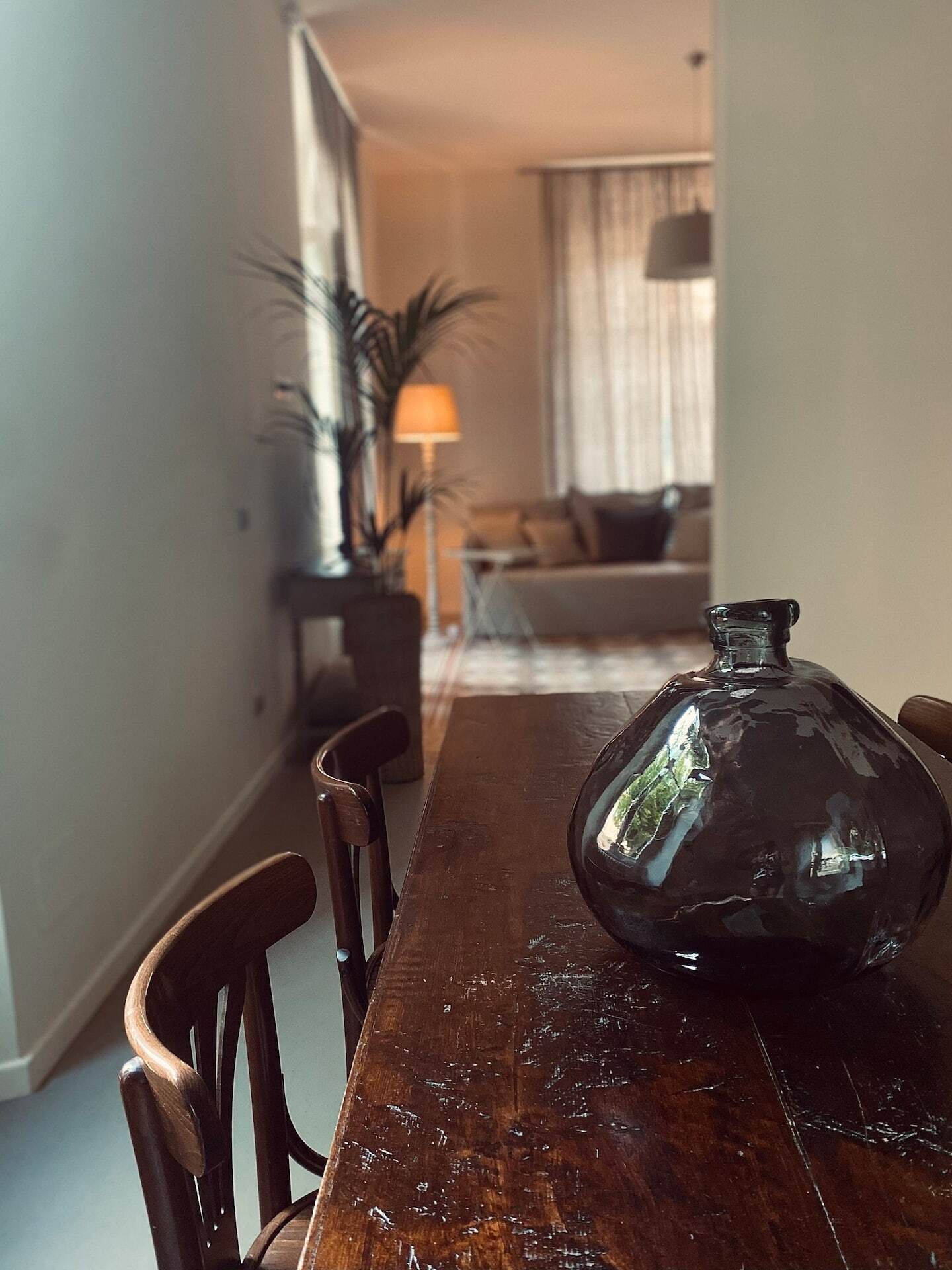 Et spisebord med stole, en stor glasvase og en lampe i en stue med sofa og gardiner.