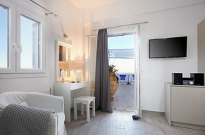 A hotel room with a bed, a desk, a chair, a TV, and a view of the sea. - Milos Villas (Santorini)