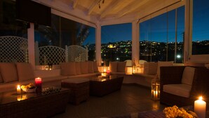 A cozy lounge area with wicker furniture, candles, and a view of a cityscape at dusk. - Milos Villas (Santorini)