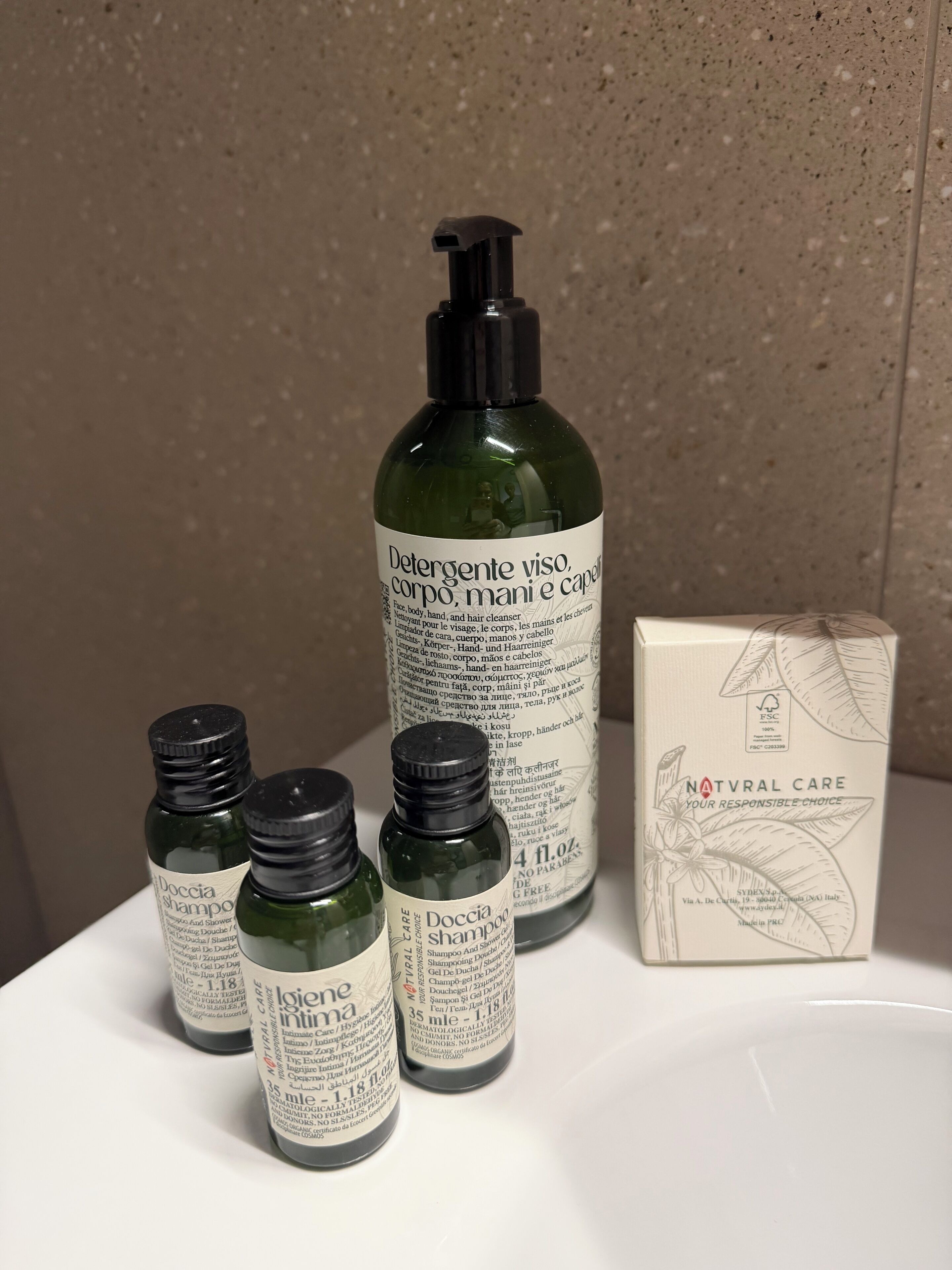 A bathroom with a white sink, a green bottle of shampoo, a white box labeled 'NATURAL CARE,' and four smaller bottles of 'Doccia Shampoo.'