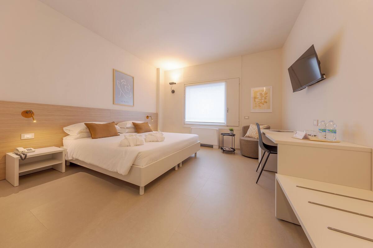 A modern hotel room with a bed, bedside table, lamp, TV, and a desk with a chair.