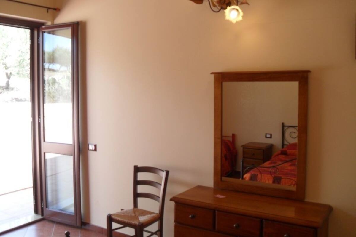 A room with a wooden dresser, a chair, a large mirror, and a sliding glass door leading to an outdoor area.
