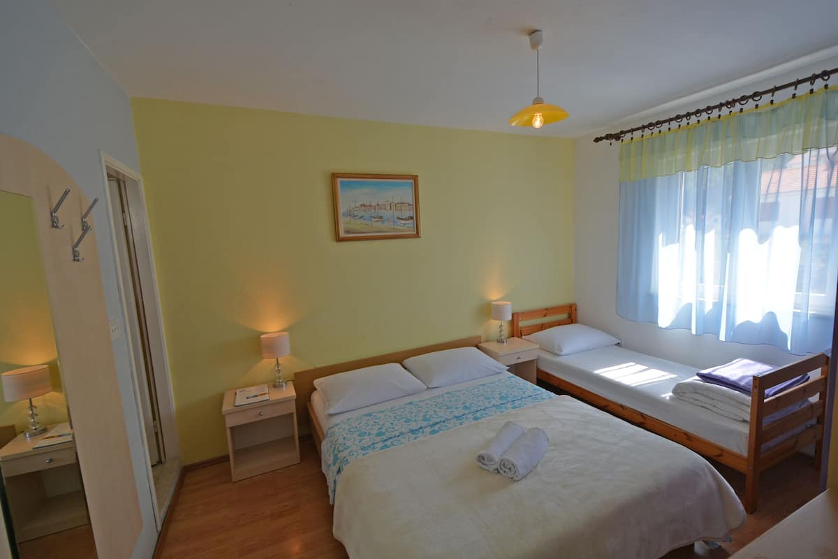 Een kamer met twee eenpersoonsbedden, een houten hoofdbord, een nachtkastje met een lamp, en een raam met dunne gordijnen. Er hangt een ingelijste foto aan de muur.