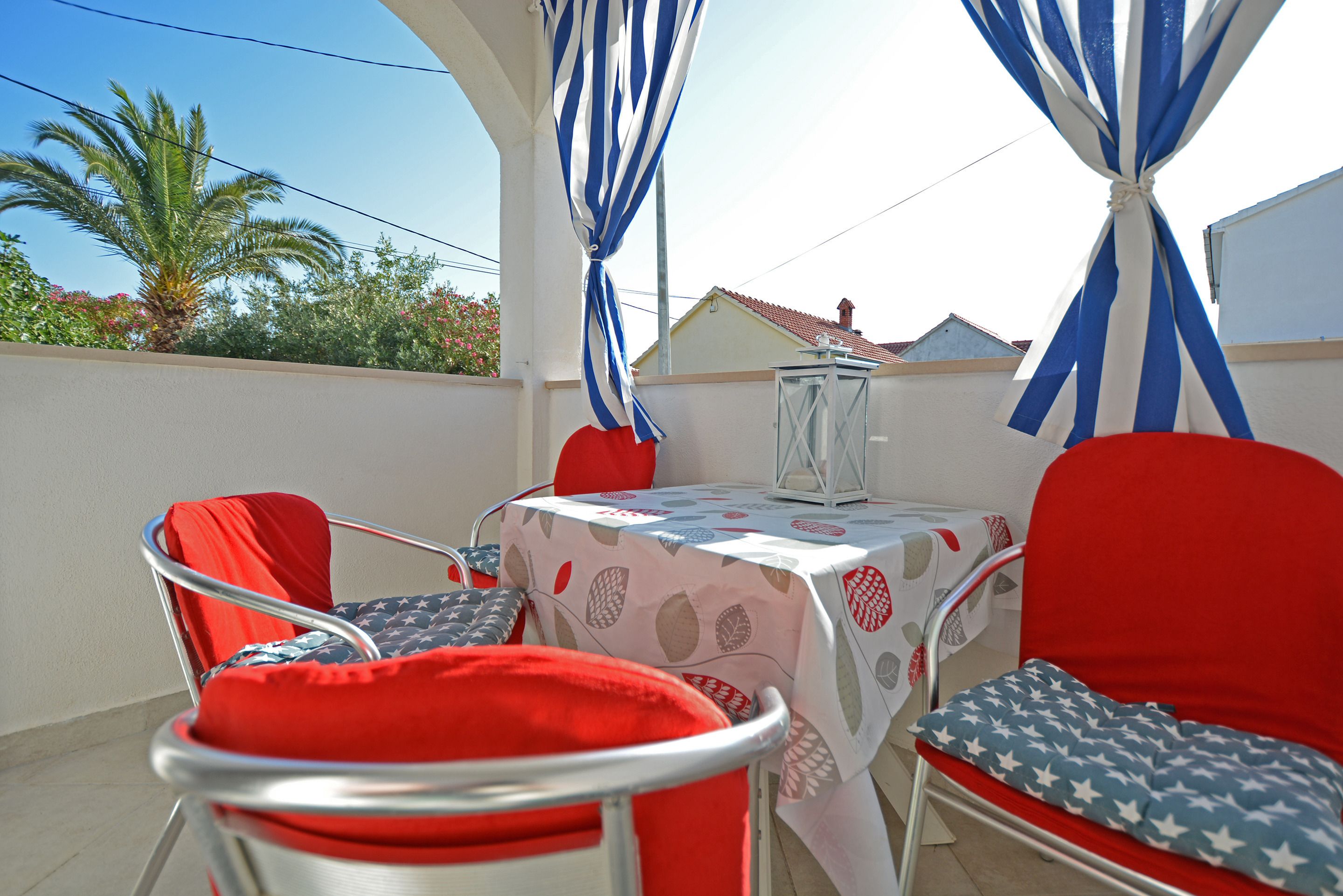 Een terras met een tafel en stoelen, overdekt met een wit tafelkleed met een bladmotief, en blauw-witte gestreepte gordijnen.