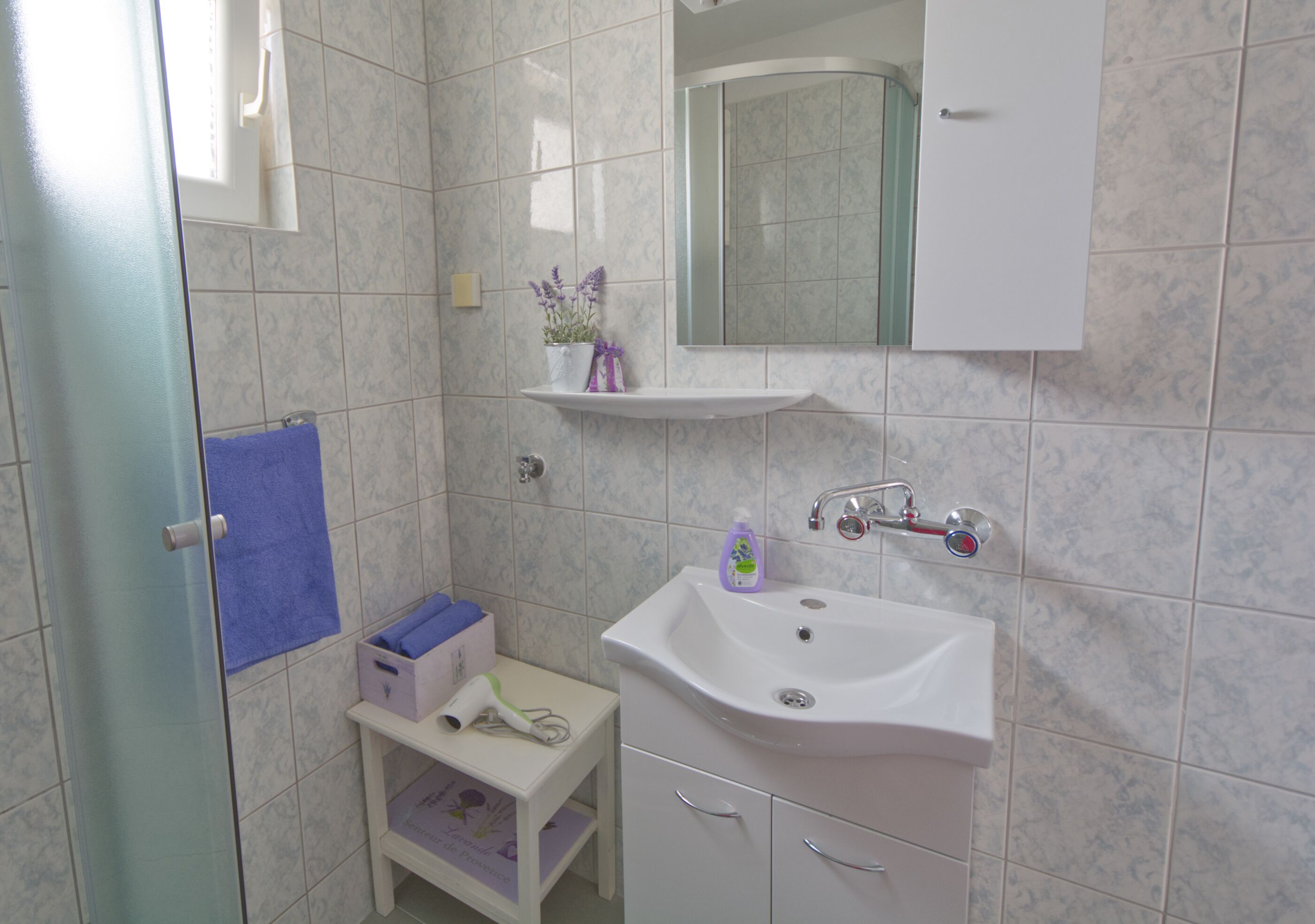 A bathroom with a white sink, a mirror above it, and a shelf with a purple bottle and a plant.