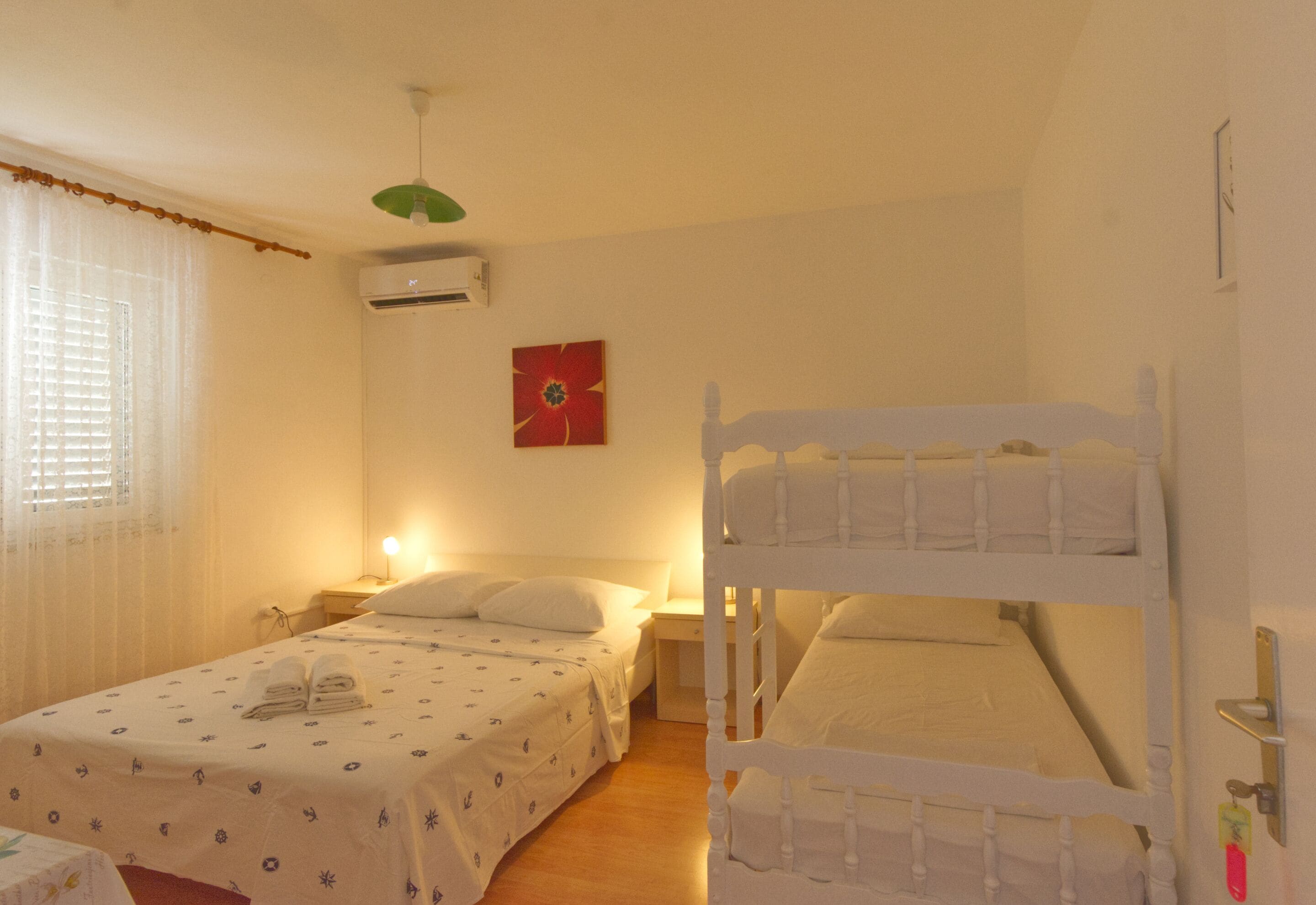 A hotel room with two beds, one with a bunk, a bedside table with a lamp, a wall-mounted air conditioner, and a window with curtains.