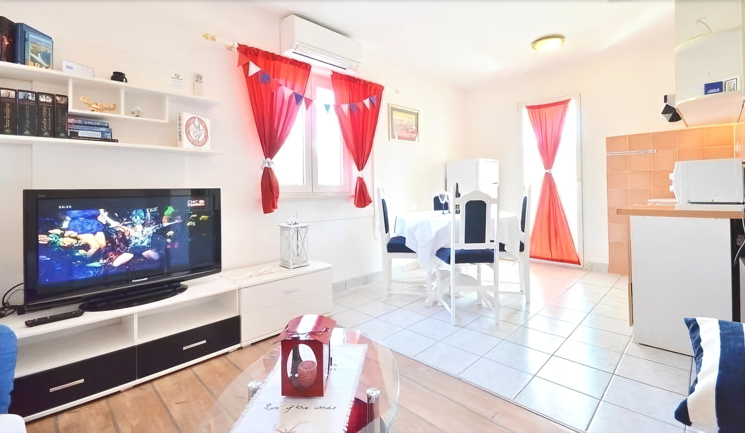 A modern living room with a flat-screen TV, a dining area with a white table and chairs, and red curtains.