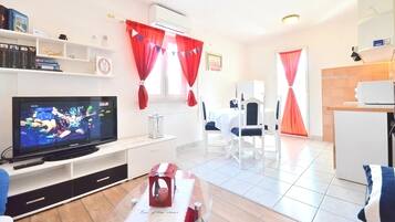 A modern living room with a flat-screen TV, a dining area with a white table and chairs, and red curtains.